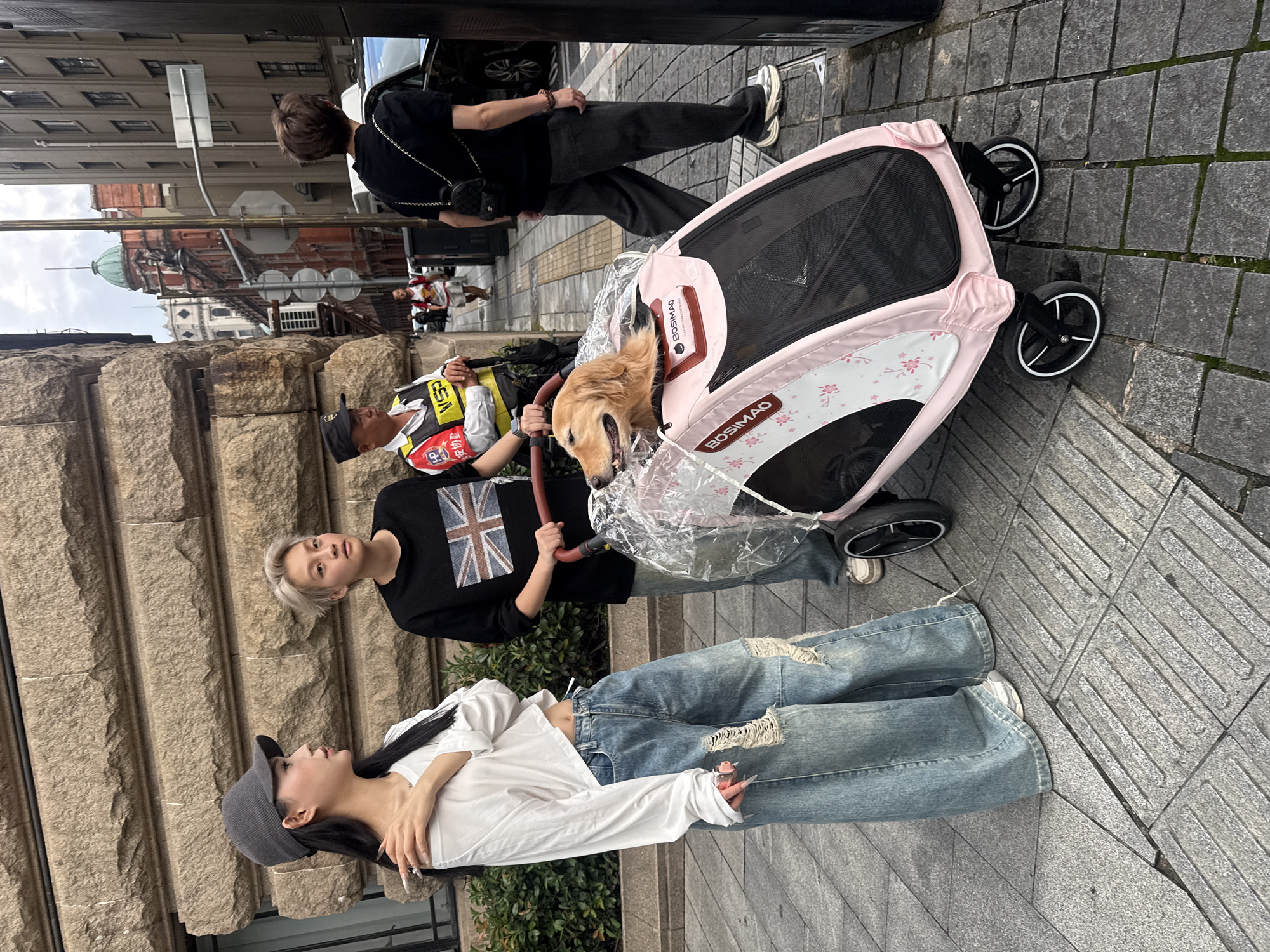In Shanghai, China, on a pedestrian sidewalk next to a cobblestone street, a young woman wearing a white long-sleeved off-the-shoulder top, ripped wide-leg denim jeans, and a grey baseball cap stands to the left. Beside her, another individual with short blonde hair, wearing a black t-shirt with a British flag graphic, pushes a pink dog stroller. A golden retriever sits in the stroller, looking out. In the background, a security guard in a yellow vest stands near a building. Further down the street, an ornate red building with a green dome is visible, along with other pedestrians and vehicles. The scene appears to be an everyday urban setting.