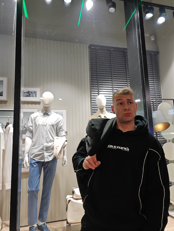 A male individual is depicted looking leftward with a slightly downturned mouth, positioned in front of a retail storefront in Shumen, Bulgaria. He is fair-skinned, with short, light-colored hair and wears a black hooded sweatshirt featuring white text "DYSTOPIA" and white piping details. A dark, possibly padded, object is slung over his left shoulder. His left arm is bent, with his hand visible near his chest. The reflection of green and white overhead lights, along with minor smudges, is visible on the glass separating the male from the store interior.
Inside the store, a clothing display is visible. On the left, a light-colored mannequin wears a patterned button-down shirt with rolled-up sleeves, light blue trousers, and blue shoes, holding a beige garment draped over its left arm. Behind it, a rack of white shirts is partially visible. Further right, a second mannequin, partially obscured, wears a light top. The background consists of a vertically paneled wall displaying framed black-and-white photographs. A dark Venetian blind covers a window or opening in the upper right. Lighting fixtures are mounted on the ceiling. A decorative lamp with circular elements is visible on the far right, and a light-colored bag with brown accents rests on the floor.