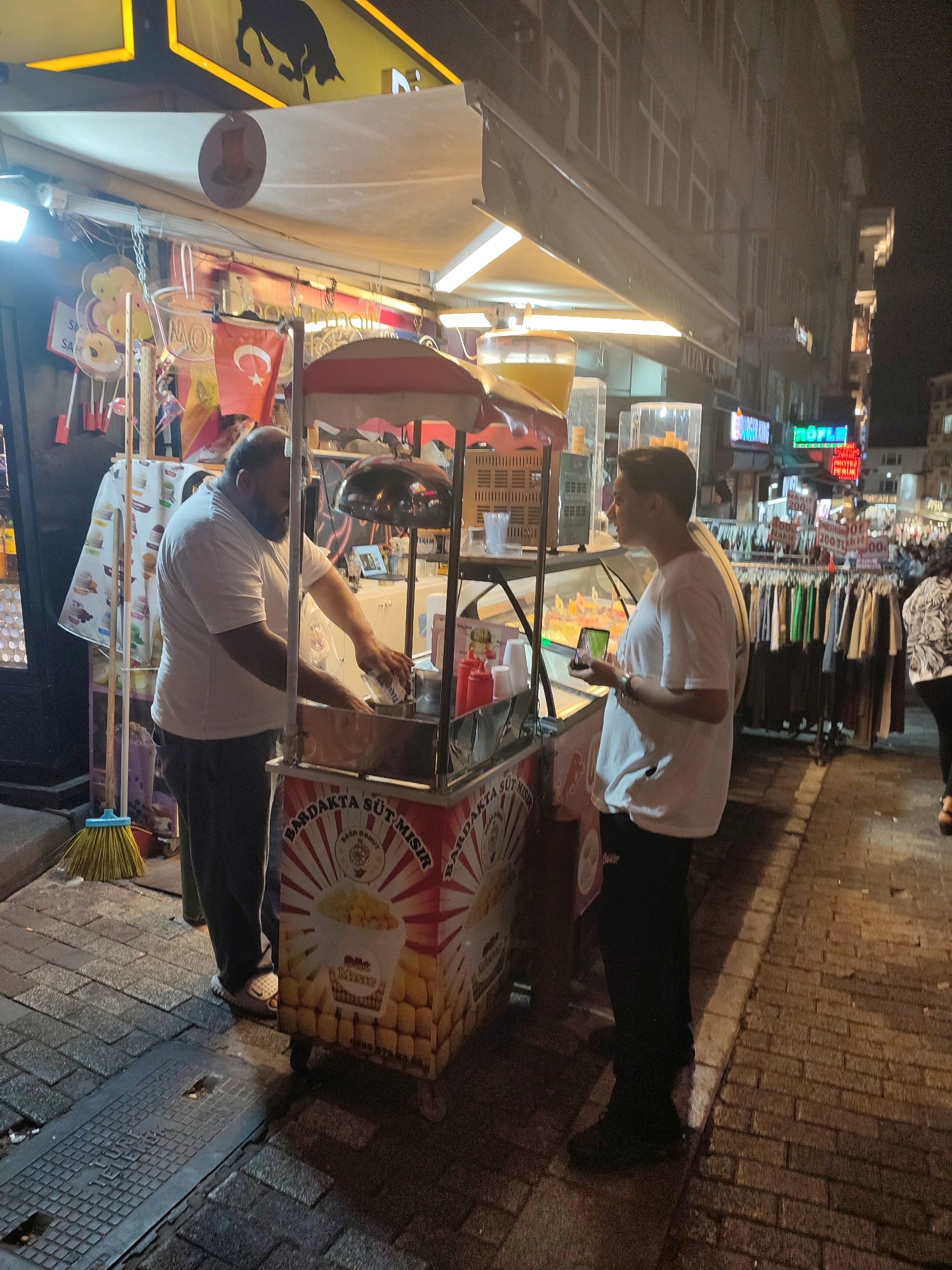 In Kadıköy, Turkey, a man stands behind a food cart, preparing a treat for a customer. The cart, decorated with images of corn, displays the text "BARDAKTA SÜT MISIR" and "BOĞAZİÇİ DONUT". The man is wearing a white t-shirt and dark pants, and is actively working with a bag of food. In front of the cart, a young man in a white t-shirt and black pants holds a smartphone and observes the preparation. The scene takes place on a brick sidewalk at night, with a street and buildings visible in the background, including neon signs and clothing racks. A broom leans against the cart, and a display of hanging items, possibly toys or snacks, is visible on the left.