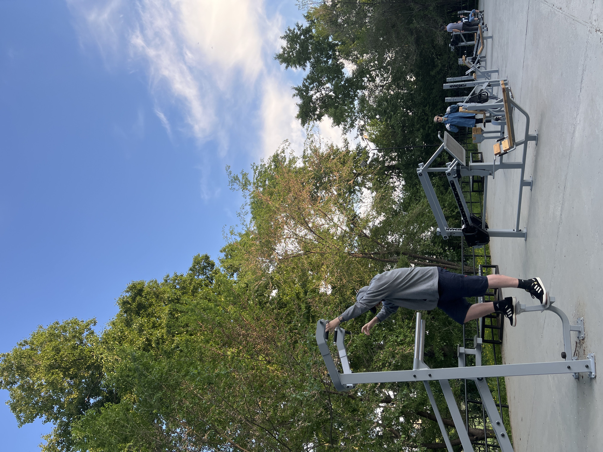 The image depicts an outdoor gym located in a park in Khabarovsk, Russia. Several individuals are present, engaged in various exercises on different pieces of equipment. In the foreground, a person in a gray hoodie and dark shorts is stepping onto a leg press machine, gripping a bar for support. Further in the background, a person in a blue jacket is standing near a chest press machine, and other individuals can be seen using various weight machines. The gym area is paved with concrete, surrounded by lush green trees and a clear blue sky with scattered white clouds. The setting appears to be a public space designed for physical fitness, with multiple workout stations arranged in a row.