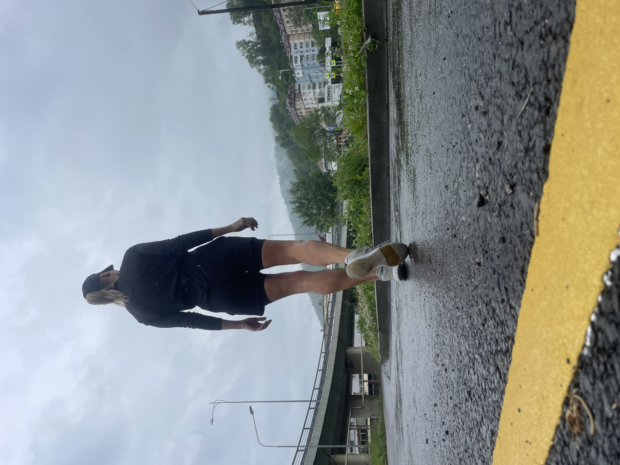 A woman stands on wet pavement next to a yellow painted line. She is wearing a black long-sleeved shirt, black shorts, a black cap, and light-colored sneakers. A bridge is visible in the background, along with buildings set against a wooded hillside shrouded in mist. The city of Sochi, Russia is discernible in the background. The sky is overcast and gray.