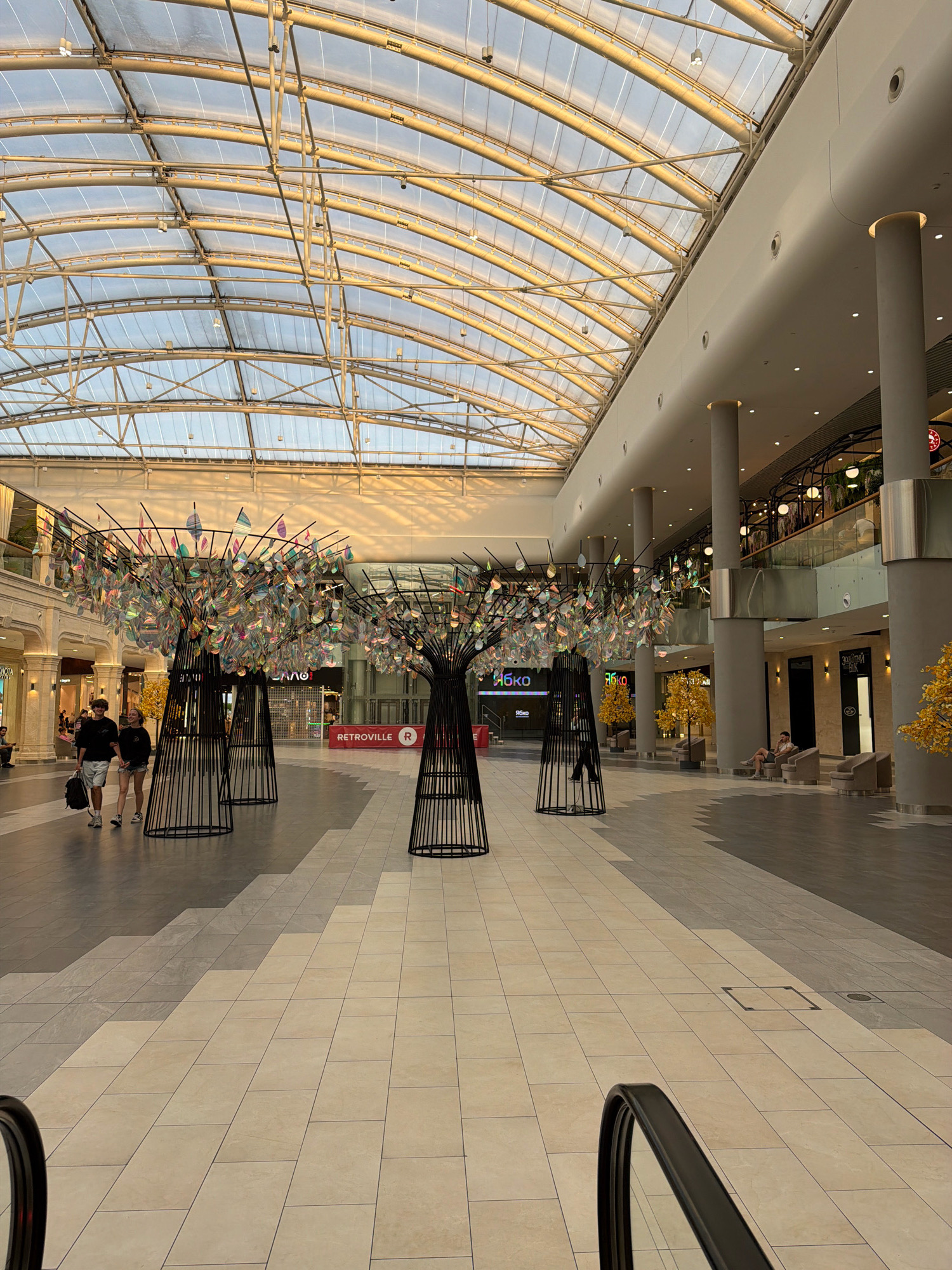 An interior view of the RETROVILLE shopping mall in Kyiv, Ukraine, taken during the daytime. The scene is dominated by the mall's architecture, featuring a large, arched glass roof that allows natural light to illuminate the space. The ceiling structure is a complex network of metal beams and translucent panels. In the foreground, the polished floor is tiled in a pattern of light and dark grey and beige squares. Several large, black, cage-like sculptures resembling trees are placed strategically throughout the open concourse, each adorned with numerous iridescent, leaf-shaped decorations.
In the middle ground, two individuals, a male and a female, are walking from left to right. The male is wearing a dark t-shirt, shorts, and sneakers, carrying a dark backpack. The female is wearing a dark t-shirt, shorts, and sneakers. Behind them, on the left, a storefront with vertical blinds is visible. Further back, on the right side of the concourse, is a row of large, grey, cylindrical columns supporting the upper levels. Various shops are visible on the upper levels, with signage indicating stores like "ЯБКО" and "RETROVILLE". Some decorative yellow foliage trees are placed along the right side of the concourse. In the far right of the image, a person is seated on a light-colored armchair. An escalator handrail is visible in the bottom left and bottom right corners of the image. The overall atmosphere is bright and modern, with the golden hues of the sunlight creating warm tones across the scene.