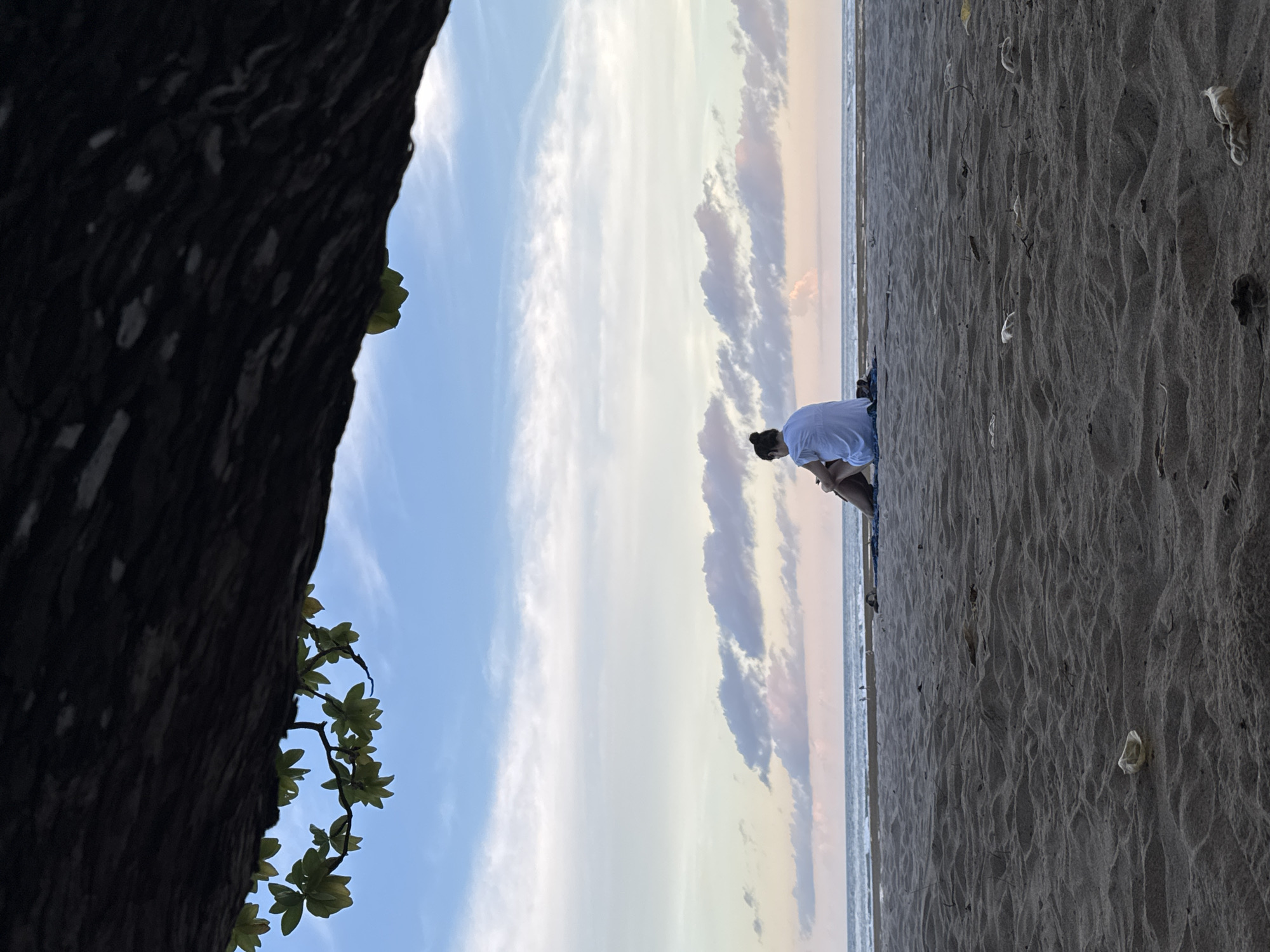 A full shot, taken from a low angle, depicts a person sitting on a sandy beach in Nusa Dua, Indonesia, under a partly cloudy sky. To the left is a large, dark tree trunk with visible green leaves. The beach stretches horizontally, covered with scattered debris. The person, dressed in a white shirt and dark bottoms, is positioned in the center of the frame, gazing at the horizon. The sea is visible in the distance between the clouds and the sand.