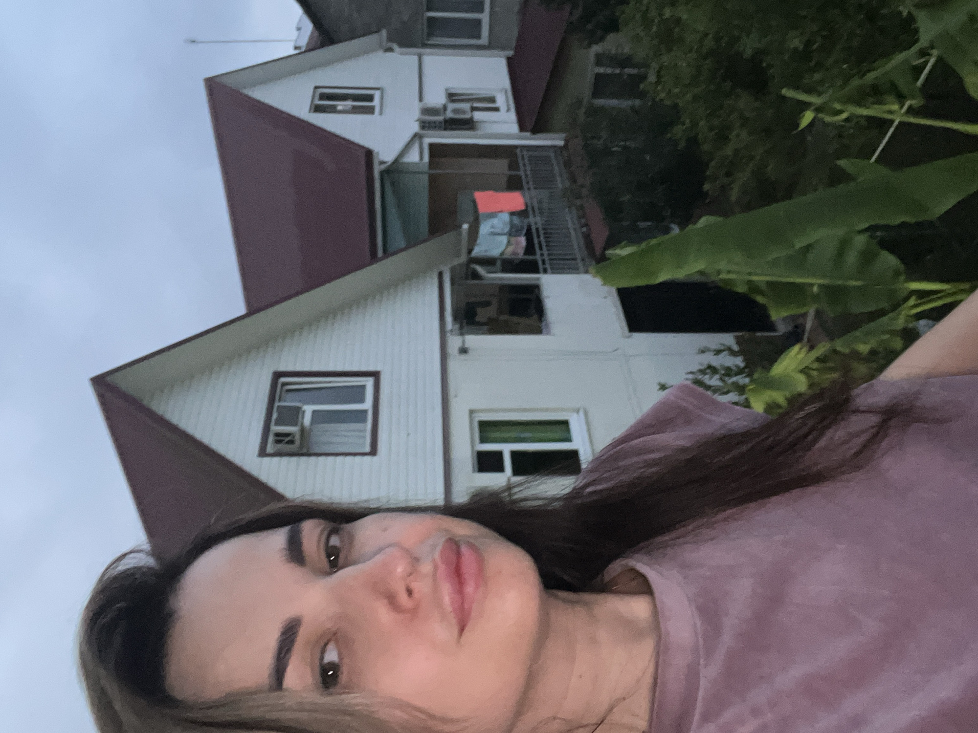 A woman is taking a selfie outdoors in front of two houses. The woman is in the foreground, facing the camera and slightly to the left, wearing a pink t-shirt and her long, dark hair is visible. Behind her, two white houses with dark red roofs are visible. The house on the left has a window with an air conditioning unit. The house on the right has a balcony with a railing and clothes hanging on it, one piece of which is a bright pink rectangle. There are also large green leaves visible in the foreground to the right of the woman. The sky is overcast.