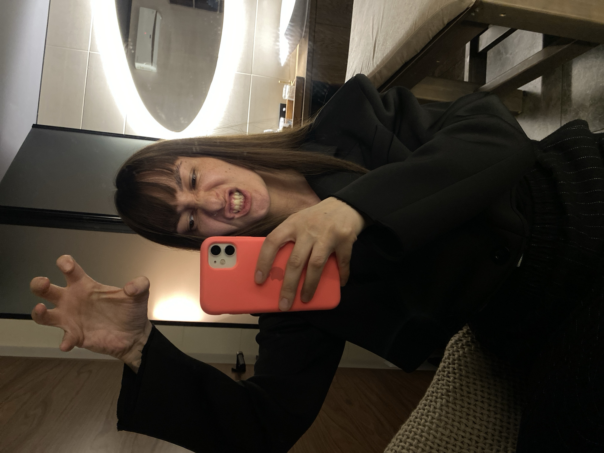 The media file is a mirror selfie, rotated 90 degrees clockwise. It features a person, likely a young to middle-aged female, with long dark brown hair and bangs, wearing a dark (possibly black) blazer or jacket. She is holding a coral-pink smartphone with a visible Apple logo outline in her right hand, using it to capture her reflection.
The person is actively posing with a playful or exaggerated "monster" expression, baring her teeth, wrinkling her nose, and widening her eyes. Her left hand is splayed out with bent fingers, mimicking a claw. A small hoop piercing is visible in her left nostril.
The setting appears to be an indoor space, possibly a modern bathroom or dressing area in Votkinsk, Russia. A large mirror with an integrated circular light strip provides bright illumination for the scene. In the background reflection, parts of a light-tiled wall, dark countertop, a glimpse of a sink faucet, and glass containers on a wooden shelf are visible. To the right, a wooden structure (possibly furniture legs) and textured fabric (perhaps a blanket or upholstery) can be seen. The floor visible in the reflection appears to be light-colored wood or laminate. No legible text is present in the image.
