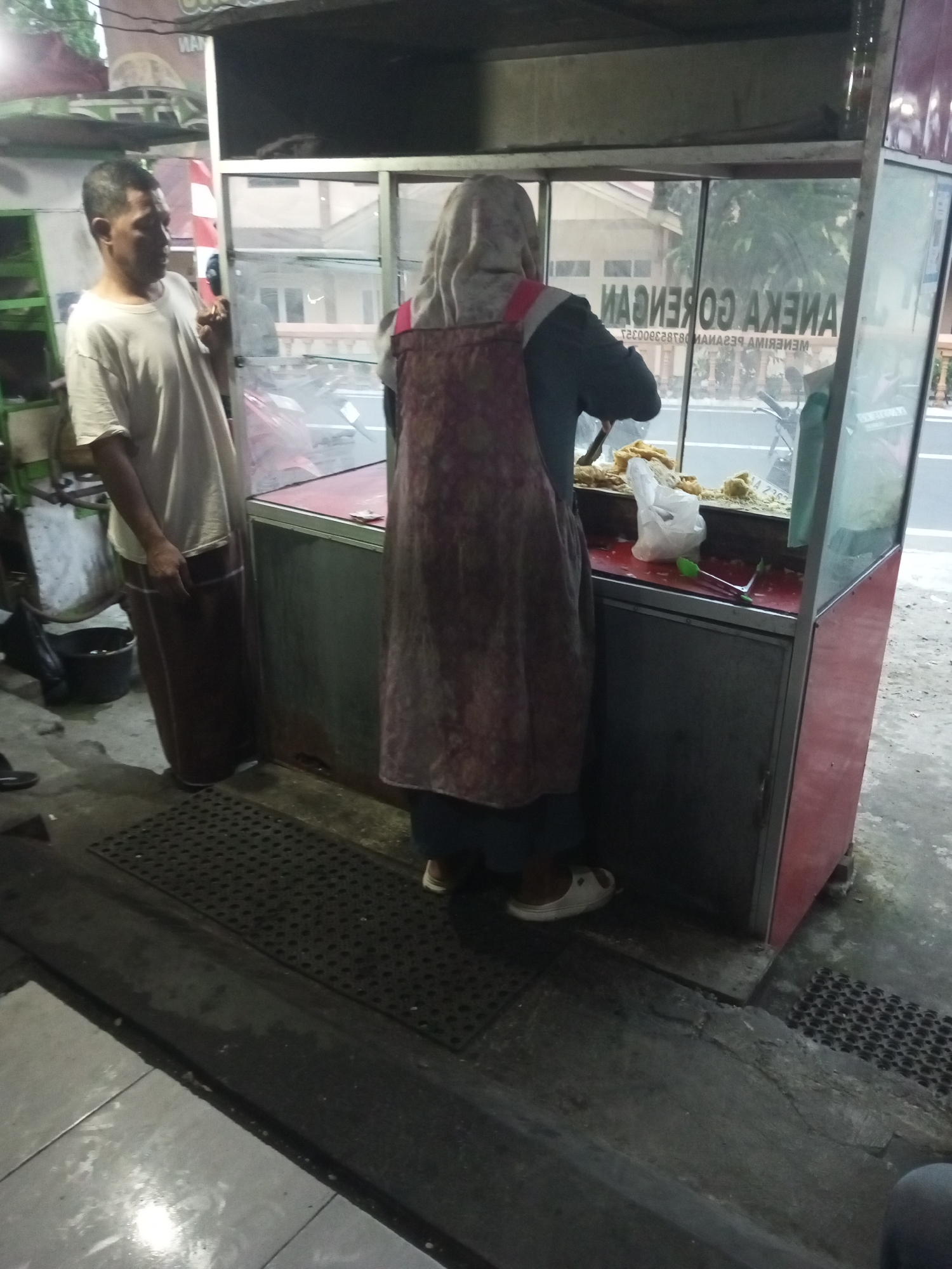 In Andongsili, Indonesia, an outdoor food stall operates with a man in a white t-shirt and a sarong standing to the left, facing a food vendor. The vendor, wearing a headscarf and an apron, is preparing food behind a glass counter. The stall features a red and gray structure with shelves displaying food items. A white plastic bag and a pair of green tongs are visible on the counter. The ground is a mix of concrete and tiled surfaces, with a dark textured mat in front of the stall. In the background, a road with a parked motorcycle is visible, alongside buildings and trees.