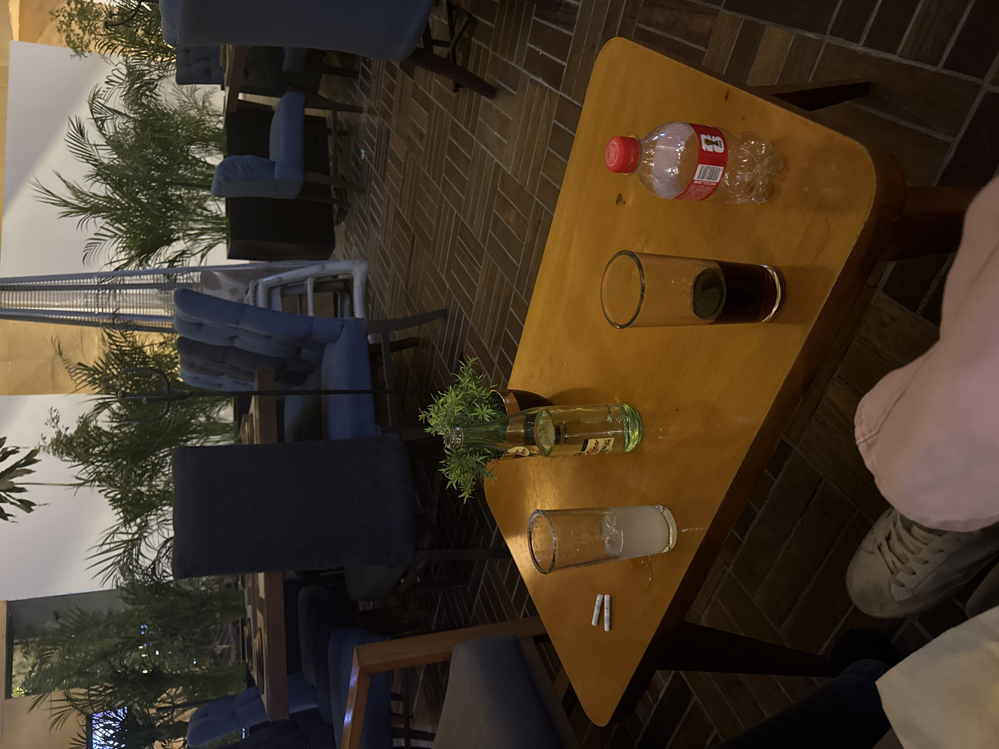 The image shows an outdoor seating area in what appears to be a cafe or restaurant. The tables and chairs are occupied by people who are partially visible. There are plants and greenery nearby. On a table, there is a bottle of Coca-Cola, a glass of liquid, a vase with greenery, a glass of what appears to be water, and two cigarettes. The location is possibly in Puebla, Mexico, but this is based on the user request, not visual evidence.