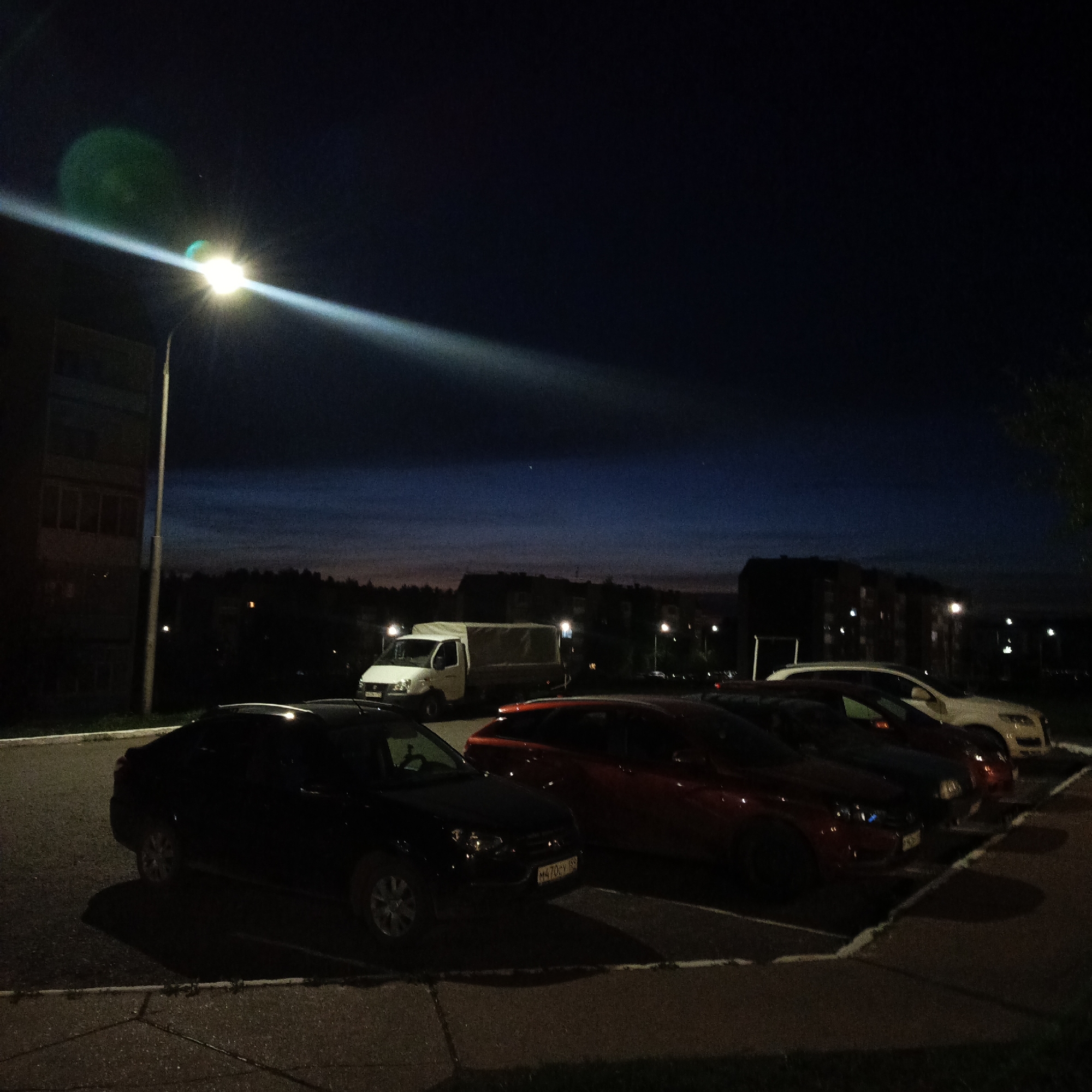 The scene is a nighttime parking lot in what appears to be an urban or suburban setting in or near the city of Марковский, Russia. The time of day is likely dusk or night, as evidenced by the dark sky and the use of artificial lighting.
The parking lot contains several vehicles, including a white van, a black car, a red car, and what appears to be a light-colored SUV. The cars are parked in a row.
A street light is illuminated, casting a bright glow across the scene.
In the background, there are buildings, possibly apartment buildings. There are also trees and other vegetation, suggesting the parking lot is located in a residential area.
The overall impression is of a quiet, ordinary scene.