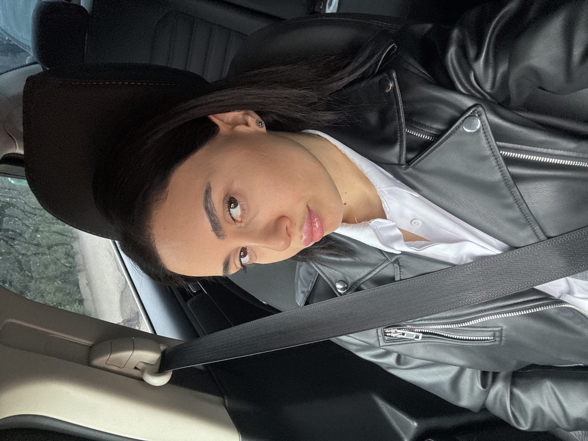 A young woman is sitting in the passenger seat of a car. She is wearing a white shirt under a black leather jacket and has on her seatbelt. She has dark hair and light brown eyes. The car is likely moving, given the blurred scenery visible through the car window. The location of the scene is unknown, but the request specifies that it is supposed to be in Tbilisi, Georgia.