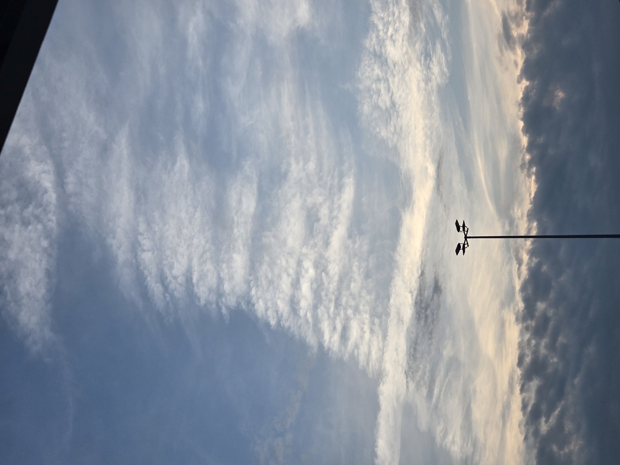 The image is a vertical, medium-shot photograph of a twilight sky with scattered clouds and a lamppost in the foreground. The sky transitions from a light blue near the horizon to a darker blue at the top. Wispy, white cirrus clouds are visible across the upper portion of the sky, with a more dense, dark gray layer of stratocumulus clouds occupying the lower third. A tall, thin lamppost with two heads is silhouetted against the sky in the right half of the frame. The top edge of the image is partially obscured by a dark, horizontal bar, suggesting it might be taken from indoors or under an overhang. The image does not provide any specific visual cues to pinpoint its location within Ansbach, Germany.