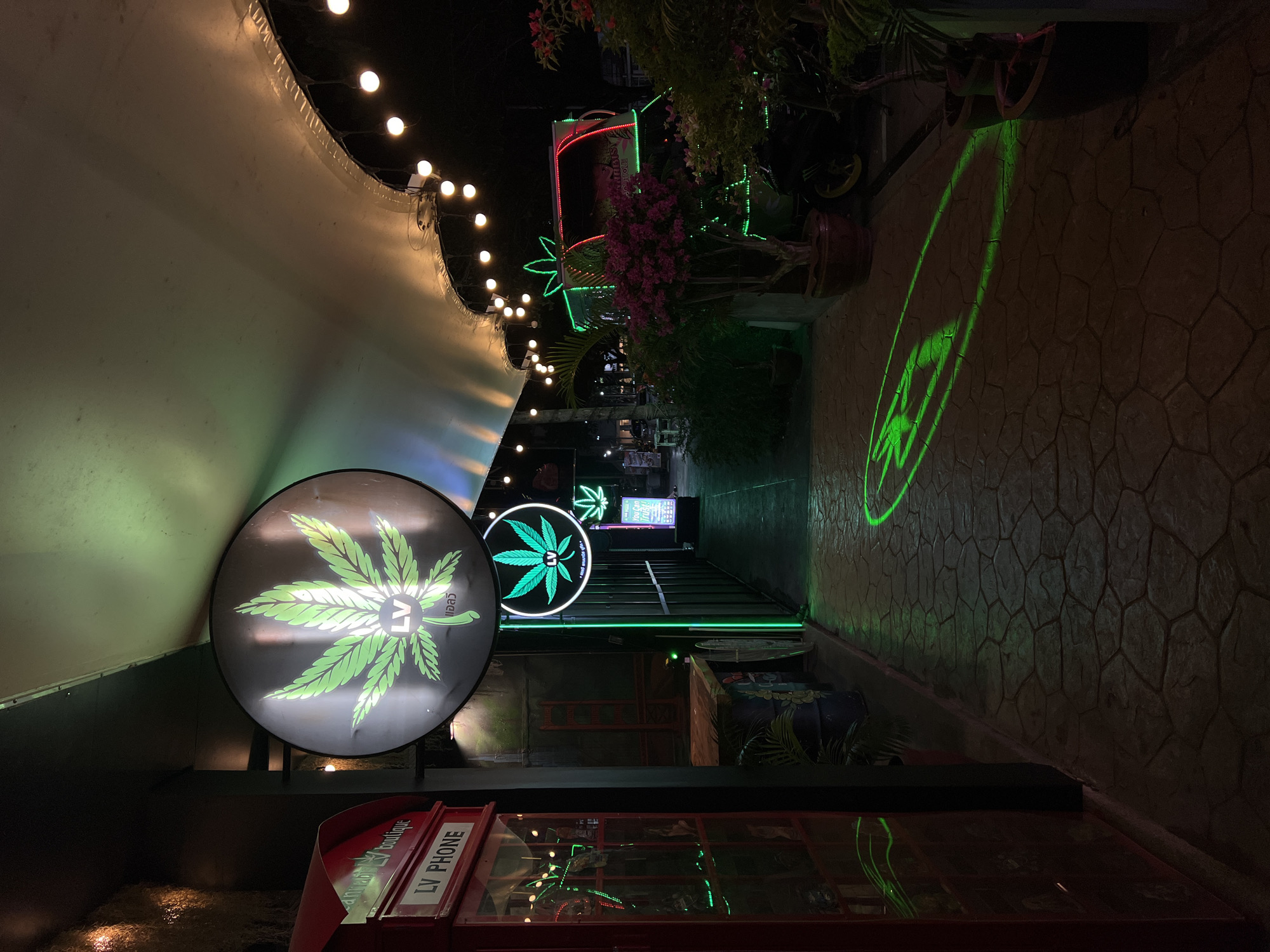 Here is a description of the image:
The image captures a narrow alleyway at night, potentially a storefront or entertainment district. The alley is illuminated by a mix of overhead string lights, neon signs featuring cannabis leaf designs, and green light projections.
A large, circular sign with a cannabis leaf logo dominates the foreground, positioned above a red phone booth. Further down the alley, more cannabis-themed neon signs are visible. Green light projects a cannabis leaf design onto the cobblestone-style paving.
Potted plants are arranged on either side of the alley, adding a touch of greenery to the urban setting. The overall ambiance suggests a cannabis-themed establishment or district.