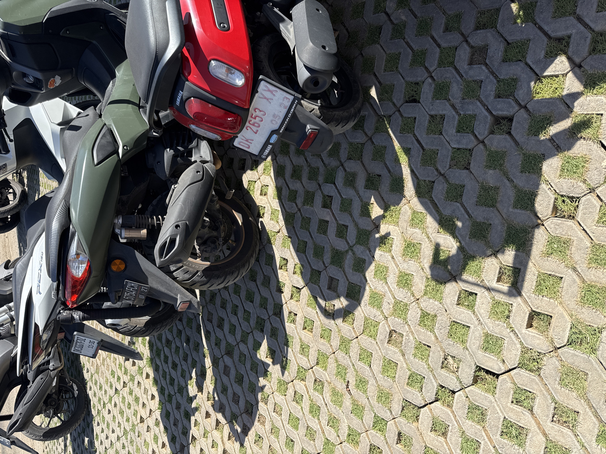 The image depicts a parking area in Kuta Selatan, Indonesia. Several motorcycles are parked on interlocking concrete pavers with grass growing in the gaps. The angle and proximity of the motorcycles suggest a crowded parking situation. License plates are visible, with numbers DK5754FCH, DKSGG7FDZ, and DK2653XX. The motorcycles are various colors (red, white, and olive green) and models, including a Yamaha. The lighting indicates a sunny day. Shadows of people (possibly a person holding a camera phone) are visible on the pavers.
