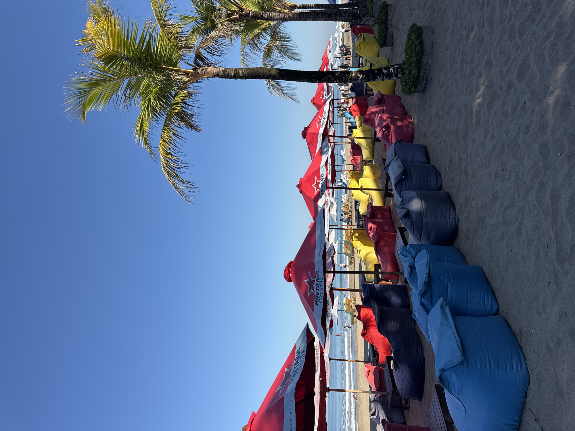 The image shows a beach scene in Kuta, Indonesia, featuring rows of colorful beanbag chairs and red umbrellas bearing the "Bintang Crystal" logo. The chairs are arranged in lines on the sand, with the colors alternating between blue, dark blue, red, and yellow. The umbrellas are positioned behind the chairs, providing shade. In the background, the ocean is visible with people lounging on beach chairs and swimming in the sea. Palm trees line the edge of the beach, with a clear blue sky above. The image appears to be taken during the day, with bright sunlight illuminating the scene.