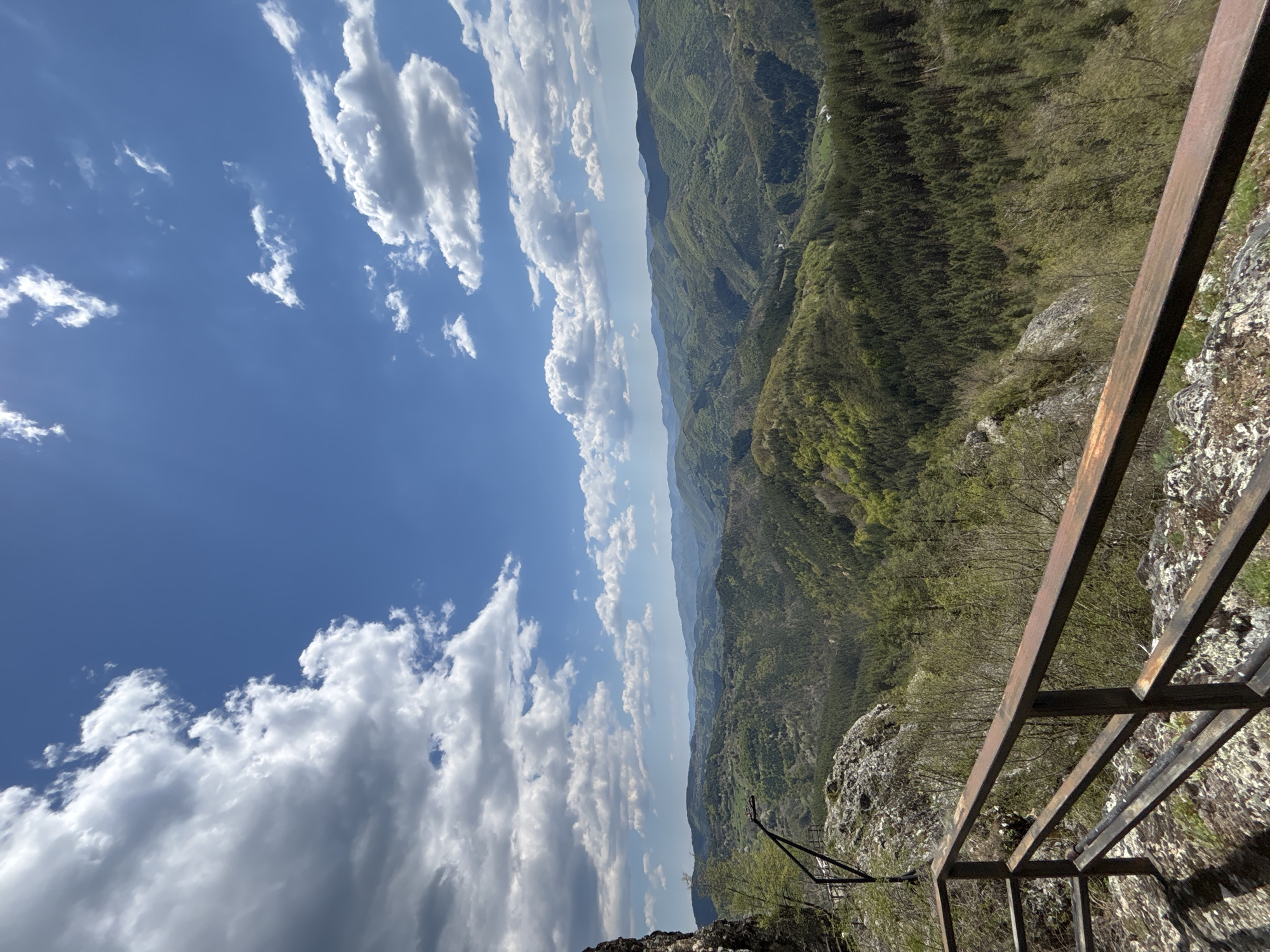 An eye-level, outdoor shot presents a landscape view from the city of Gradat, Bulgaria. The sky is filled with white clouds against a light blue backdrop. Green forested hills stretch into the distance. A rusty, metal railing forms a part of the foreground, leading down to a rocky terrain below.