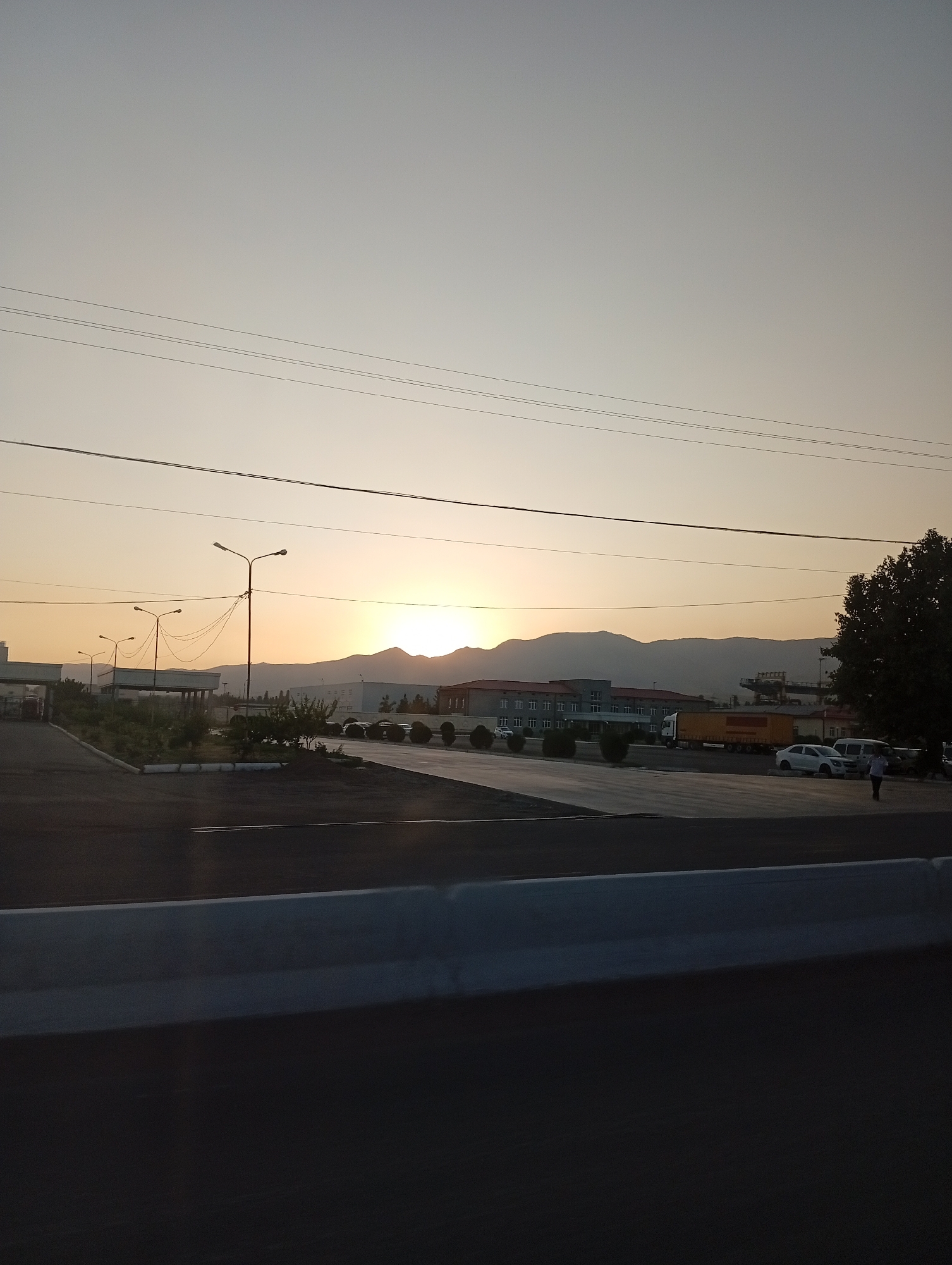 This is a wide-angle, outdoor, horizontal photograph taken at dusk in Angren, Uzbekistan. The sky is a pale, gradient of yellow and gray, with several horizontal power lines crisscrossing the upper portion of the frame. In the mid-ground, a range of subdued, dark mountains forms the backdrop. In front of the mountains, there are several low-rise buildings, one of which appears to be a factory or industrial facility with a light-colored facade and numerous windows. A large, yellow truck with a red stripe is parked in front of one of the buildings, alongside other vehicles, including white cars and a white van. Several streetlights with multiple lamps are visible, some illuminated. The foreground consists of a dark asphalt road or parking lot, with a lighter-colored, raised concrete barrier running horizontally across the bottom of the image. A single person, wearing dark clothing, is walking on the right side of the road. The overall scene depicts an industrial or commercial area at the end of the day.