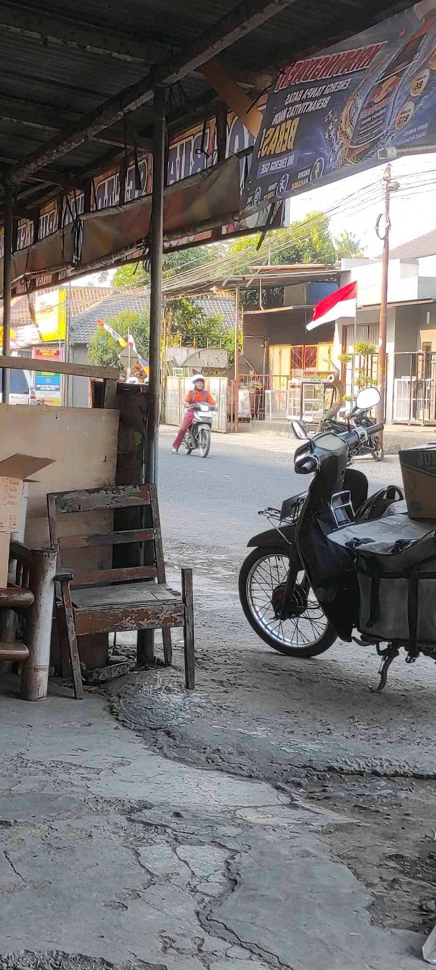 The image depicts a street scene in Purwodadi, Indonesia, likely in the late afternoon judging by the lighting. In the foreground, a weathered wooden chair is positioned on a cracked concrete surface. To the right, a dark-colored motorcycle with a large storage box on the back is parked. Beyond the motorcycle, a person wearing an orange shirt and dark pants rides a white motorcycle, facing away from the camera and towards the right of the frame. The person is wearing a white helmet. In the background, buildings with red-tiled roofs are visible, and a red and white Indonesian flag flies from a pole. The street appears to be paved, with other motorcycles and structures suggesting a commercial or residential area. The overall impression is of a typical street view in a Southeast Asian town.