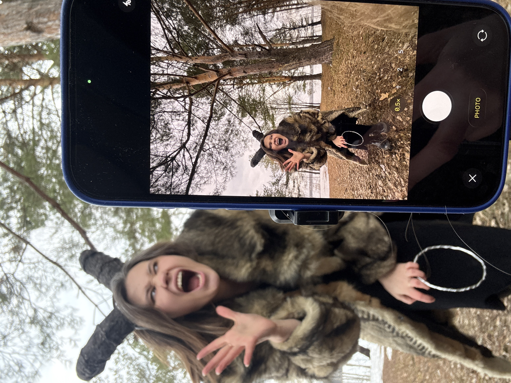 A person wearing a fur coat and horned hat is photographed using a smartphone. The background shows a forest scene with snow on the ground. The location is Chaikovsky, Russia. The person in the fur coat is making a screaming gesture with their mouth open and hands outstretched. In the phone screen image, the subject is positioned among trees with dry foliage. A second person is visible in the top-right of the frame, holding the phone.