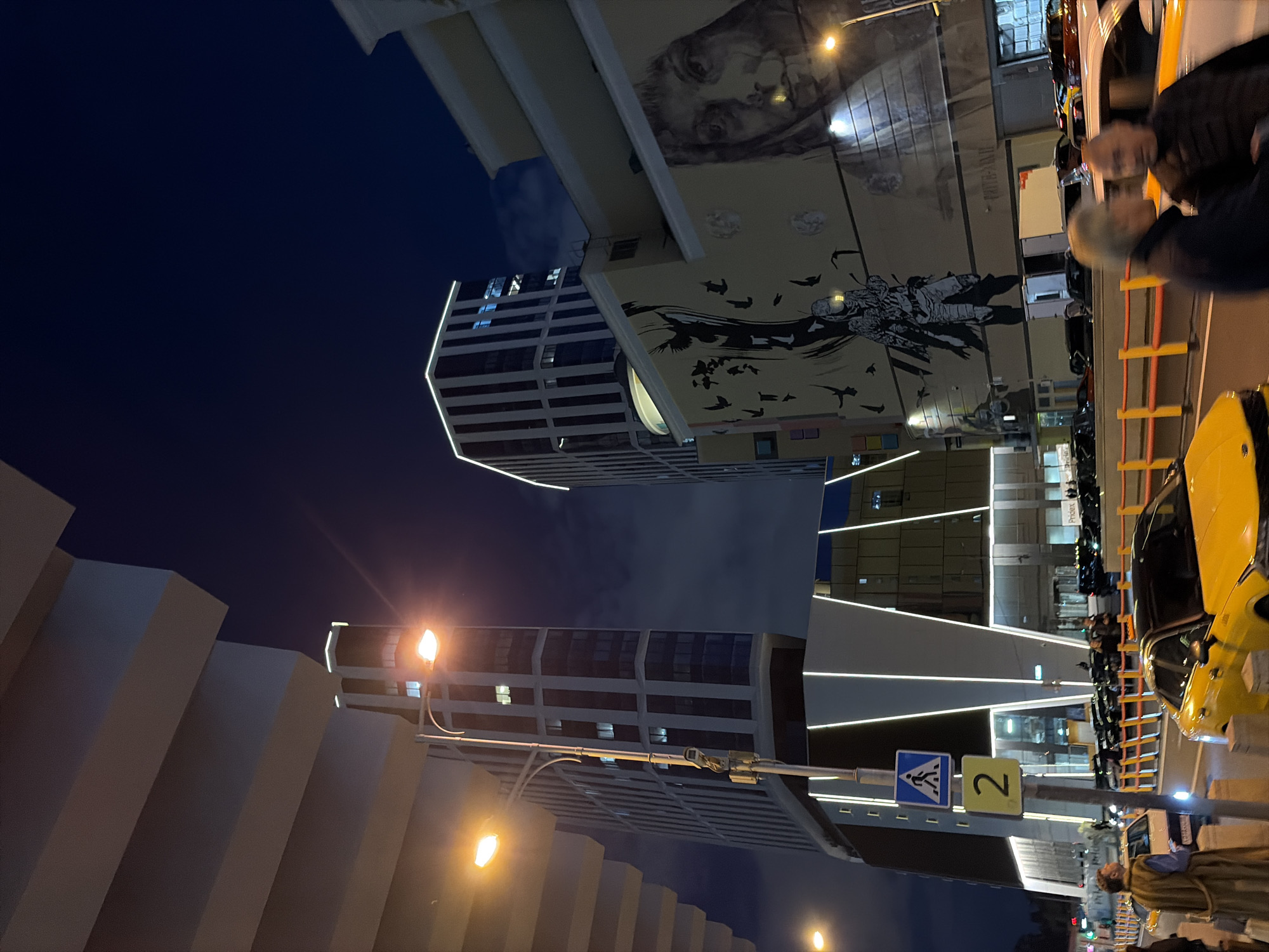 This is a medium-shot, eye-level, outdoor, vertical photograph taken at night in Moscow, Russia. The image captures a street scene with modern architecture and illuminated buildings. In the foreground, a yellow car is parked on the right side of the frame, partially obscuring another yellow car in front of it. Two men are in the foreground, facing away from the camera and talking, with one man in a dark jacket and the other in a lighter jacket. To the left of the yellow car, a person wearing a long, beige coat stands facing the street.
A street sign with a pedestrian crossing symbol and the number "2" is visible on a pole. Behind the people and cars, a brightly lit, multi-story building with a reflective facade is prominent. To the left of this building, another taller, dark-colored building with vertical stripes and illuminated window outlines stands. The facades of some buildings feature large murals, including a depiction of a face on the right and a scene with a figure and birds on the left. Streetlights illuminate the scene, casting an orange glow. The sky is dark, with some faint cloud formations visible. The street is lined with yellow and orange barriers. Several other vehicles are parked or driving on the street in the background. The overall atmosphere is urban and contemporary, characterized by artificial lighting and modern design.