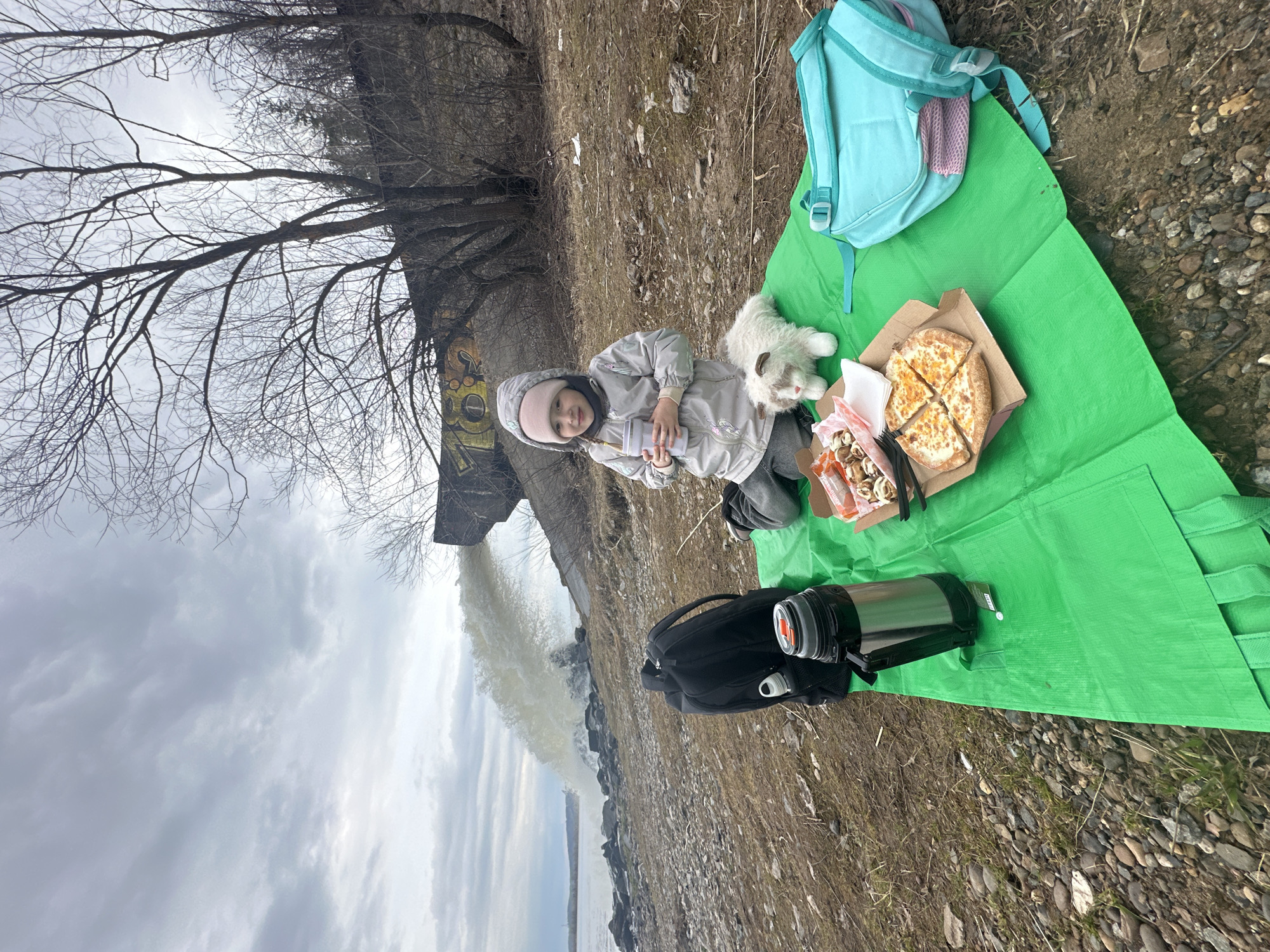 A child is seated on a green picnic blanket outdoors, next to a white stuffed animal. The child wears a gray jacket and pink hat and holds a cup. A pizza in a cardboard box and a container of food are on the blanket. A blue backpack and a black backpack with a metal thermos are also present. In the background, water is being discharged from a large pipe into a body of water under a cloudy sky. Bare trees are visible on the bank. Based on the architectural style and the presence of the industrial pipe discharging water, the location is likely in the vicinity of the industrial areas of Chaikovsky, Russia.