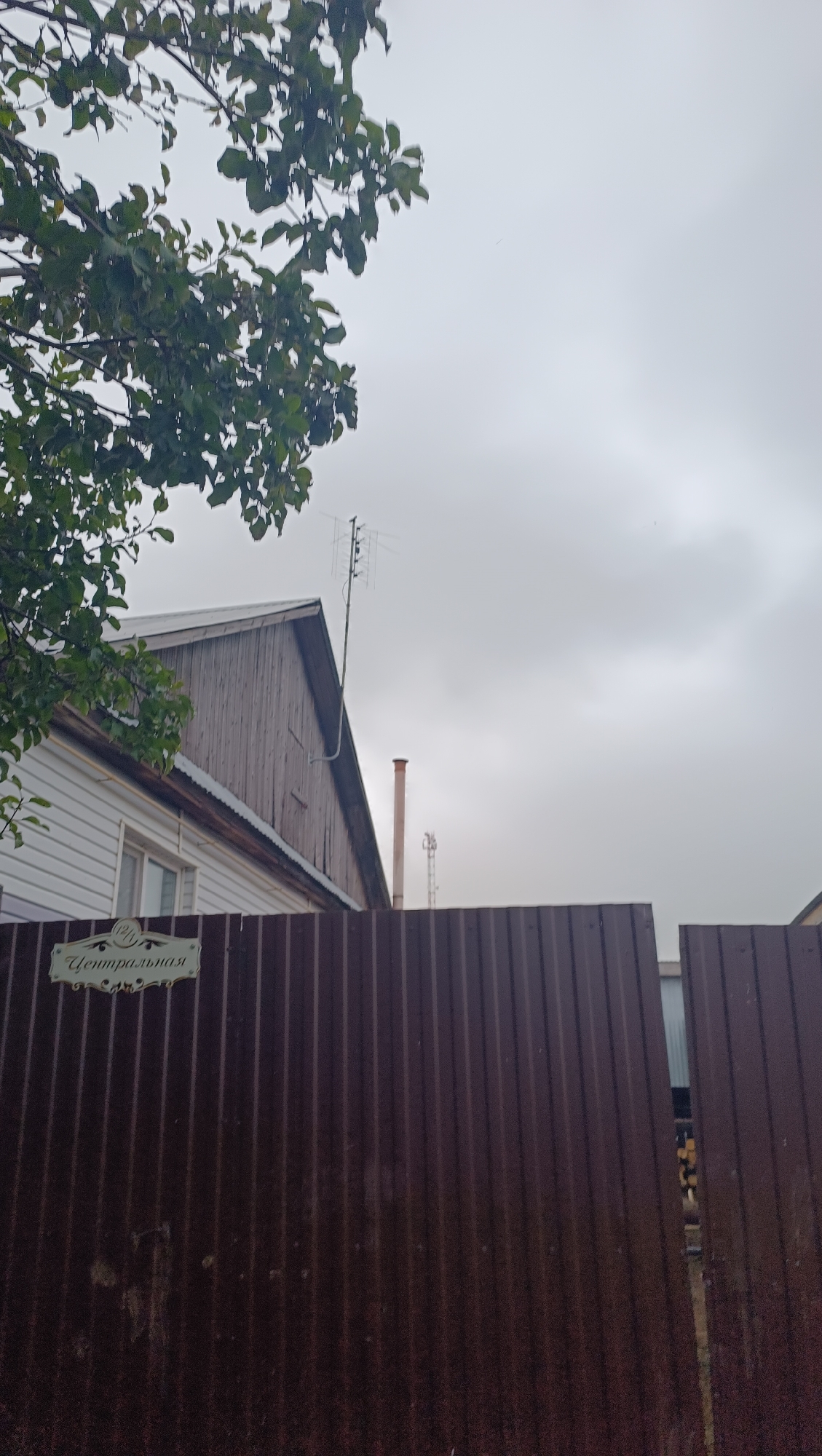 An exterior shot shows a brown corrugated metal fence in the foreground, with a sign on it that reads "Центральная". Behind the fence, a white siding house is visible, with a wooden-sided roof and a chimney. A television antenna is mounted on the roof, and a further antenna is visible in the distance. Trees with green leaves are on the left side of the image. The sky is overcast and gray. The image does not provide any specific details to determine its location in the city of Karsha, Russia.