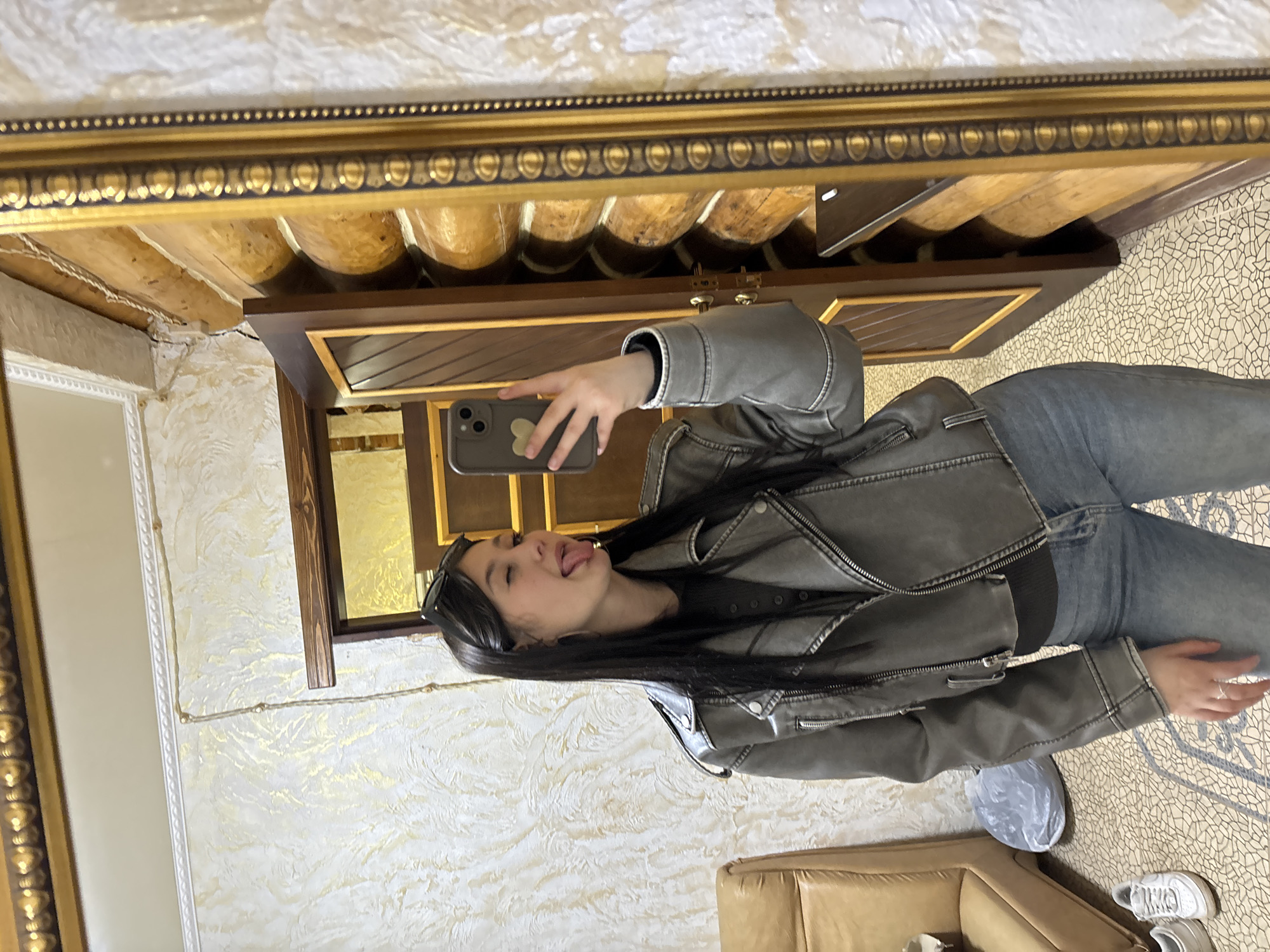 A young woman is taking a selfie in a room with textured walls and patterned flooring. She is wearing a gray jacket, dark shirt, and blue jeans. A cell phone is visible in her hand. There is a large mirror with an ornate gold frame. A brown couch is in the background. The room appears to have a rustic or log cabin aesthetic. The location is Chaikovsky, Russia.