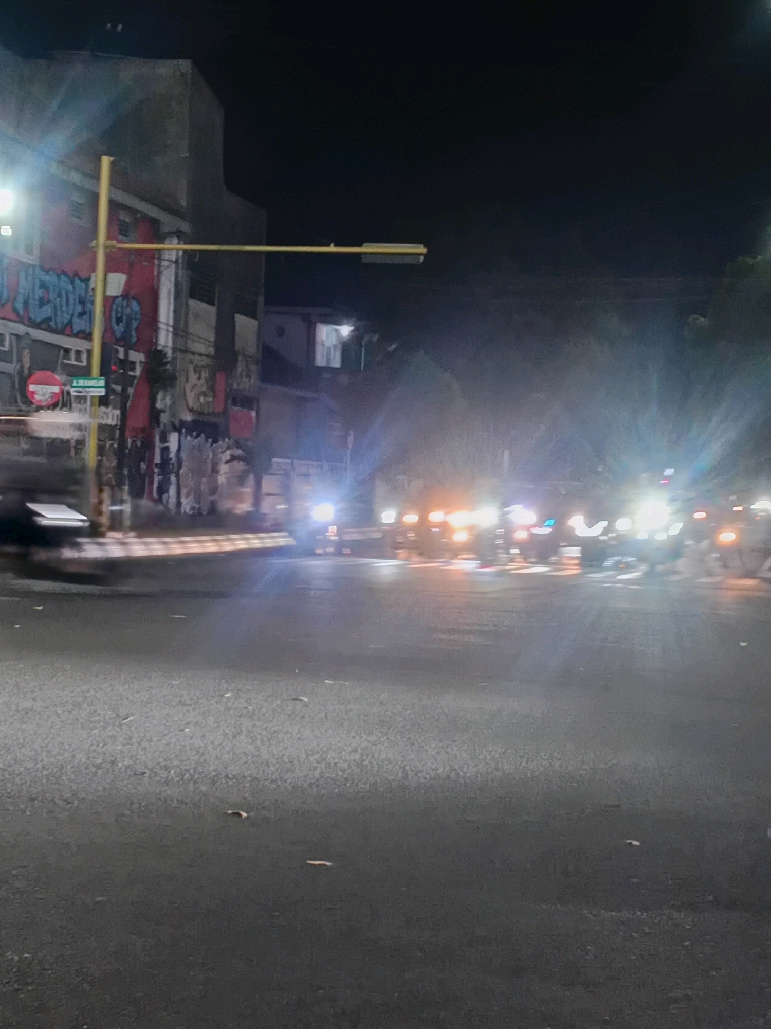 The image depicts a blurry nighttime street scene in Kudus, Indonesia. A building with graffiti, including the word "MERDEKA" in blue, is visible on the left. A yellow traffic signal pole extends across the road. Several vehicles with illuminated headlights are moving on the wet asphalt road, causing streaks of light due to motion blur. The environment appears to be an urban street.