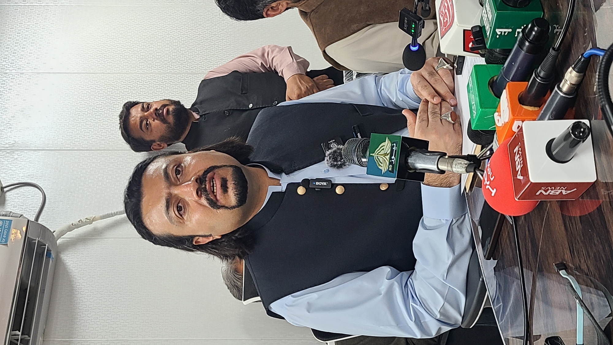 A man in a black waistcoat and light blue shirt is sitting at a table with multiple microphones from different news outlets in front of him. The logos of these news outlets are visible on the microphones. Another man is visible behind him, partially out of focus, wearing a black waistcoat over a light pink shirt. An air conditioner is visible on the wall to the left. The location may be within Quetta City Tehsil, Pakistan, but there is no direct evidence of this in the image.