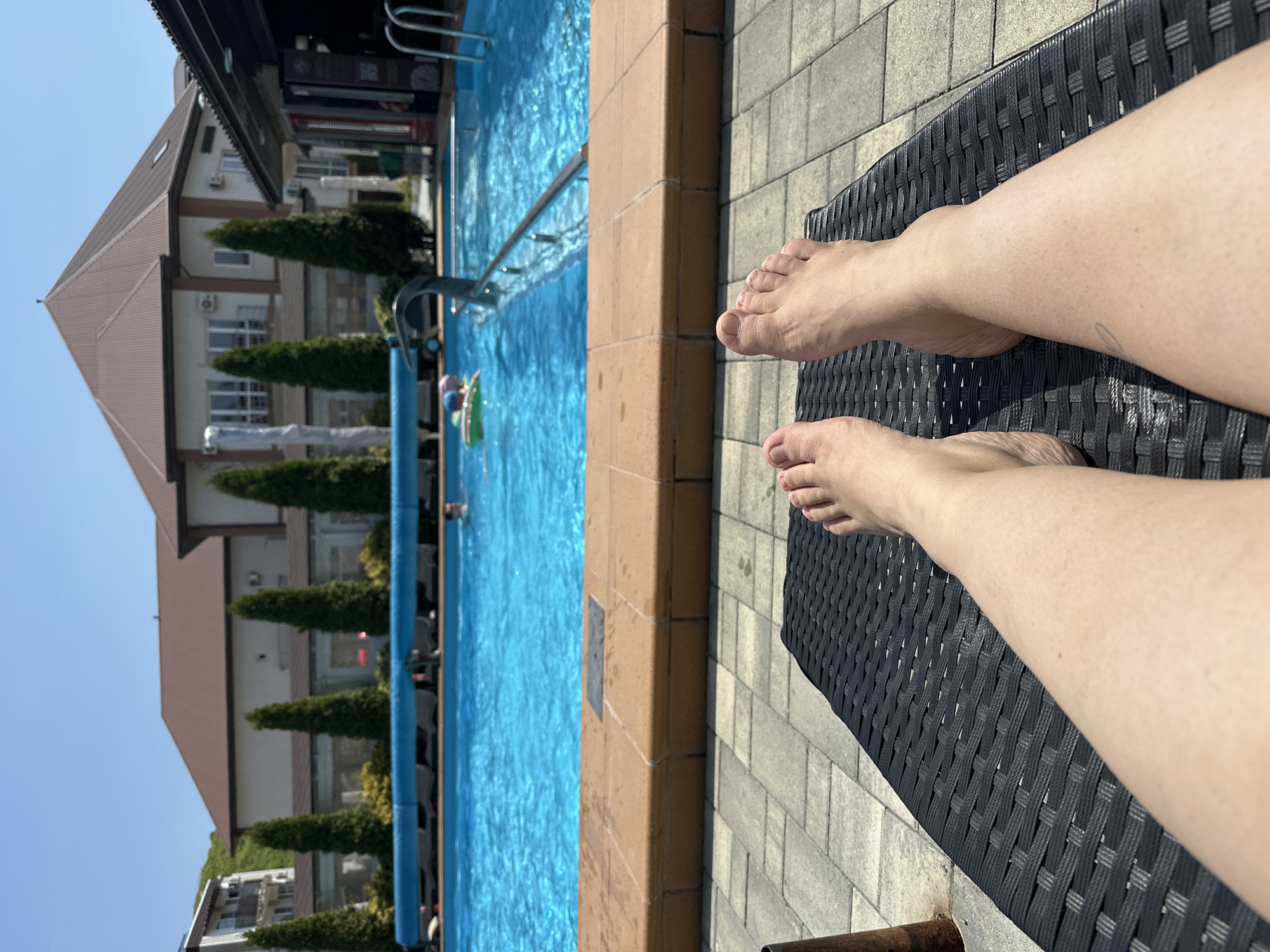 The image shows a person's legs and feet resting on a black wicker lounge chair beside a swimming pool. The pool is filled with blue water and has a brown tile edge. People are swimming in the pool. In the background is a multi-story building with a brown roof and green trees. The scene is sunlit. The image may have been taken in or near Novomikhaylovsky, Russia.