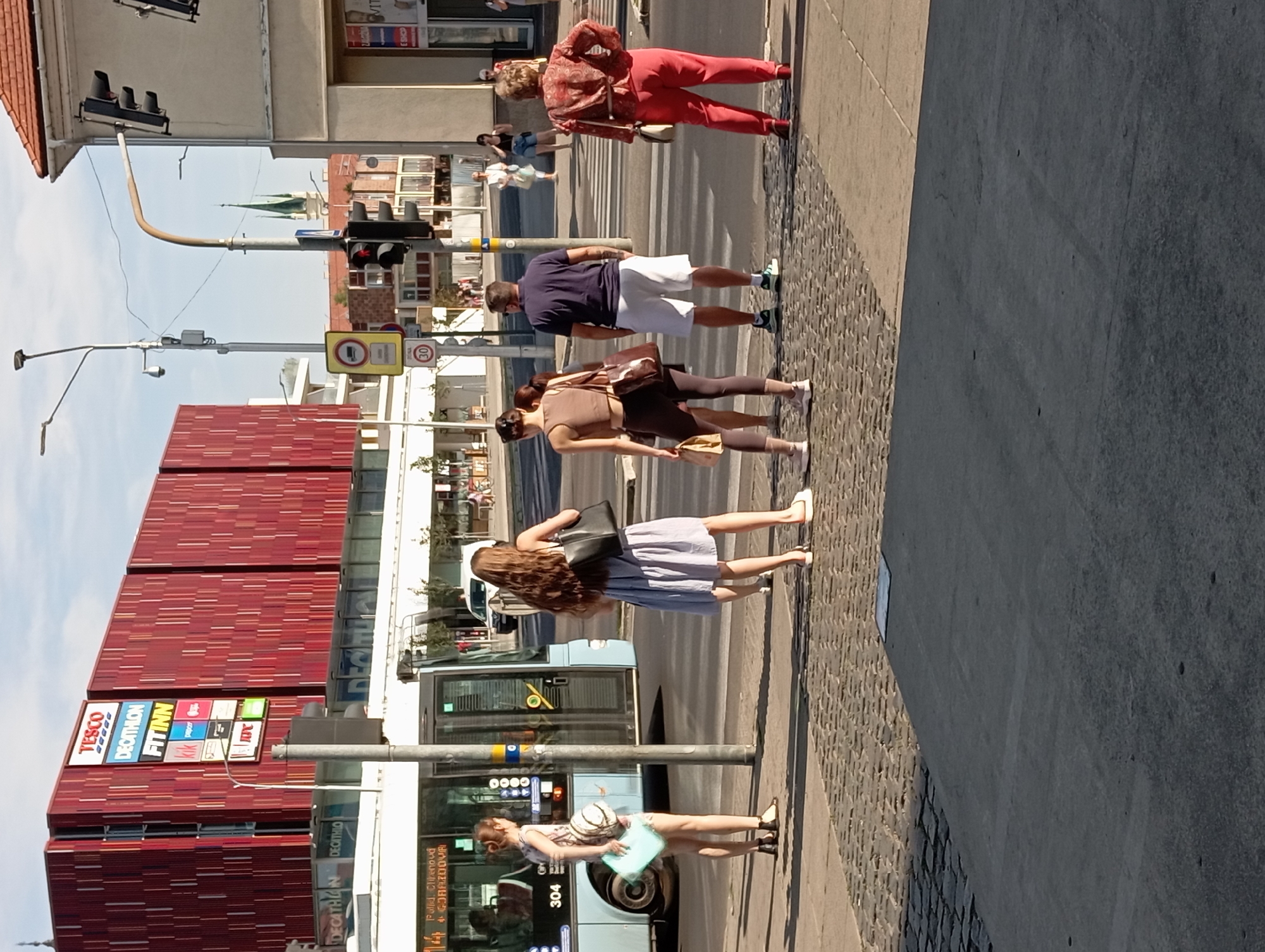 The image captures a street scene in Nitra, Slovakia, specifically at an intersection near a large red building. The building displays prominent signage for businesses including "TESCO," "DECATHLON," and "FITINN." A blue bus, numbered 304, is visible at the bus stop. Several individuals are gathered at a crosswalk, waiting to cross the street. One woman, wearing a patterned dress, stands near the bus stop, holding a light blue folder and a bag. Another woman, in a blue striped dress, stands closer to the crosswalk, carrying a black handbag. Two other individuals, a man in a blue polo shirt and white shorts, and a woman in a brown top and dark leggings, are also positioned near the crosswalk. Further down the street, another woman in a red patterned top and red pants is seen on the opposite side of the crosswalk. Traffic lights are visible, indicating the presence of a controlled intersection. The foreground is dominated by a textured cobblestone pavement. The overall impression is a typical urban setting on a sunny day.