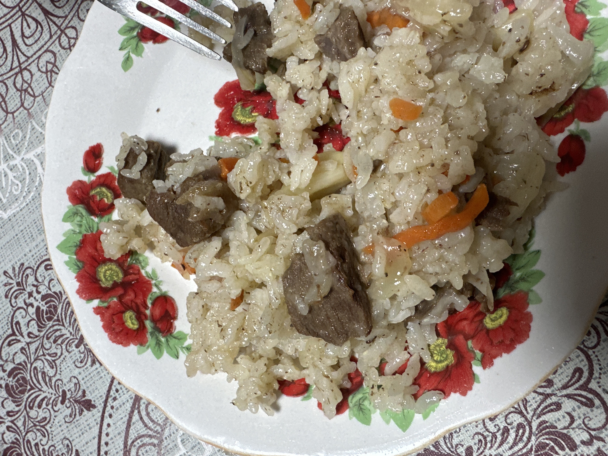 A plate of pilaf is shown in a close-up shot. The pilaf, a rice dish, contains visible chunks of meat and sliced carrots. A fork is partially visible at the top of the image, implying someone is about to eat the pilaf. The plate has a white base and is decorated with a floral pattern featuring red flowers and green leaves. The plate rests on a patterned tablecloth. The location and specific city are unknown.
