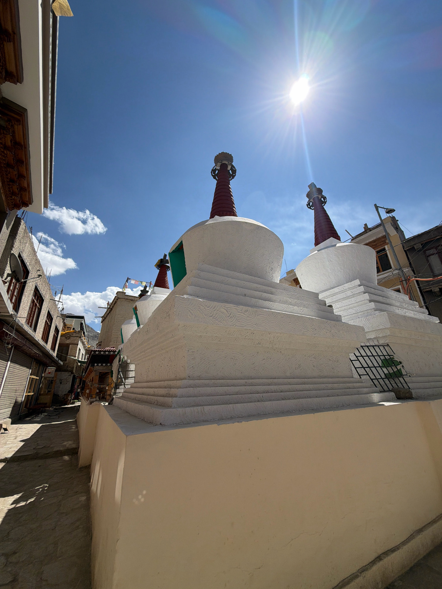 The image shows a street scene in Leh, India, dominated by a row of whitewashed stupas, a common Buddhist architectural element. The stupas have conical red tops adorned with decorative elements and are stacked in tiered layers. The foreground is occupied by the side of a stupa, with a narrow paved street visible to the left. Buildings with windows and shutters line the street, some with intricate wooden carvings on their eaves. The sky is a vibrant blue with a few scattered clouds and the sun is prominently featured, creating lens flares and casting strong shadows. The overall impression is a sun-drenched, atmospheric view of a religious and residential area in Leh.
