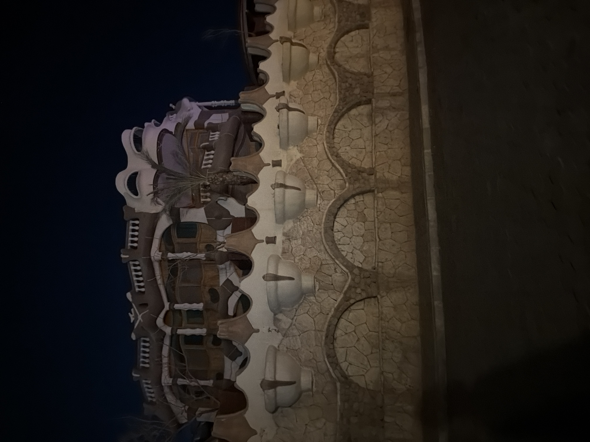The image captures a nighttime view of a distinctive building and its accompanying wall in Sharm El Sheikh, Egypt. The building, situated in the background, features an unusual, organic architectural style with rounded, protruding elements and decorative railings. Its facade is a mix of light-colored surfaces and darker, possibly wooden or textured sections. A palm frond is visible near the top right of the building.
In the foreground, a stone wall separates the property from the street. The wall is constructed from irregular, light-colored stones and is topped with a series of decorative, bowl-like structures that appear to be planters or architectural features. Below these, the wall is segmented by arched openings, also filled with the same irregular stonework. A paved street runs in front of the wall, and a dark shadow, possibly from a person, is cast on the lower left portion of the image. The overall ambiance is dark, with artificial lighting illuminating the building and wall.