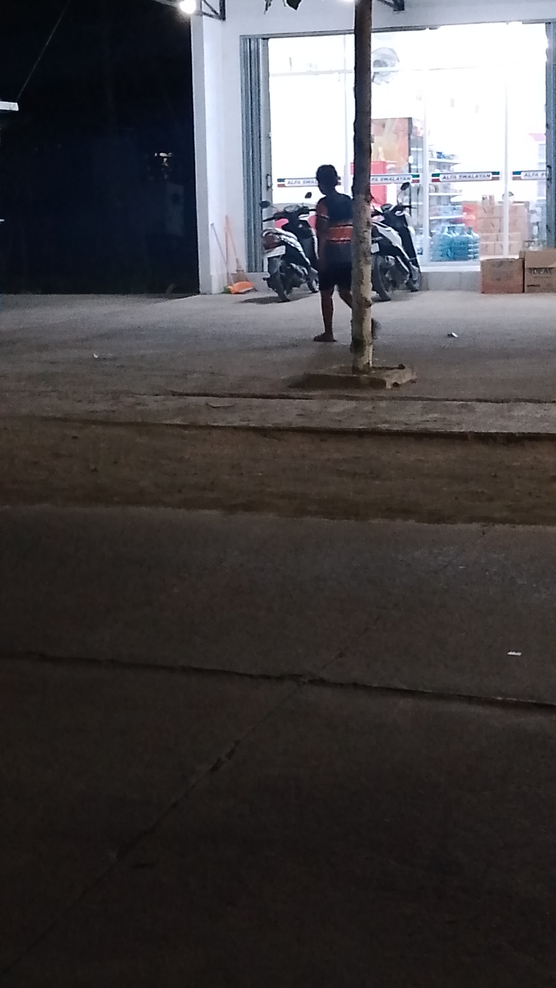 The image shows a person standing on a paved area in front of a store. The store appears to be a convenience store, with glass doors and shelves stocked with items visible through them. There are two motorcycles parked outside the store. The ground is paved with asphalt, and there is a concrete curb. A thin, dead tree trunk is standing near the person. The lighting suggests it is nighttime or late evening. The location is identified as Karanglangu, Indonesia.