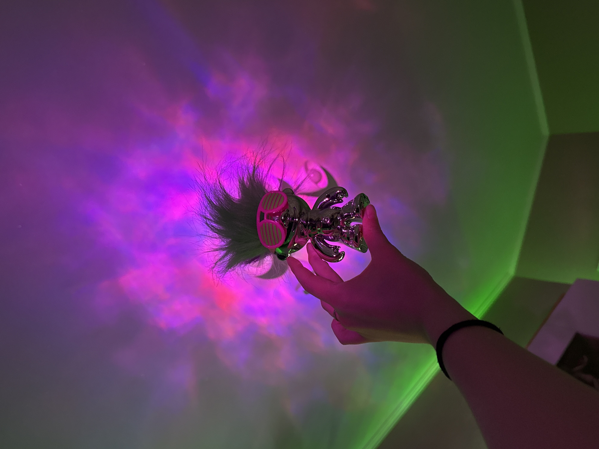 A person's arm and hand are holding a silver troll doll with pink sunglasses, with multicolored lighting patterns, including pink, purple, and green, projected onto a white ceiling. The location is unknown. The person's arm has a black wristband. A partial view of a corner of a room and a white wall with a book or framed item on it are visible in the bottom right corner of the image.