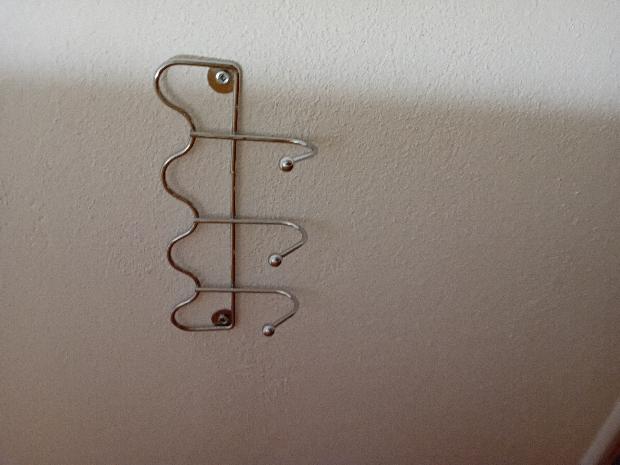 An outdoor shot shows a textured, off-white wall with a silver, four-hook rack mounted on it. The rack is curved at the top with a wave-like design. Each hook has a small, round ball at its end. There are visible screws holding the rack to the wall. The lower left of the image shows a darker area, possibly a shadow or another object. The scene appears to be in Zuberec, Slovakia, based on the context provided.