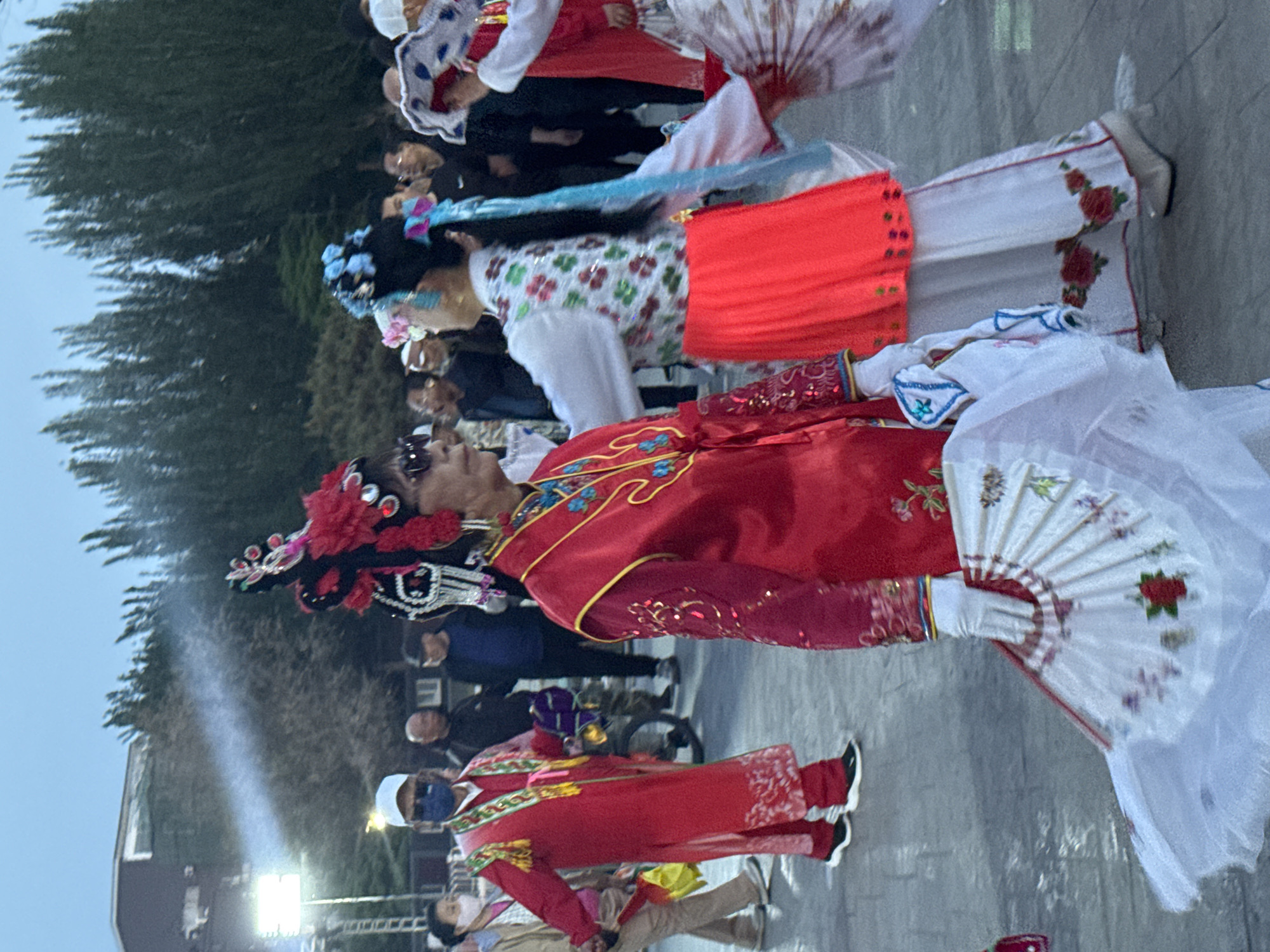 A medium shot, taken at eye level, depicts a line of people in traditional Chinese opera attire in what appears to be the city of Jinzhou, China.
The foreground features a person dressed in a vibrant red robe with intricate gold embroidery, holding a large white fan adorned with a floral pattern. Behind them is another person in a white and red dress with colorful floral patterns and a red sash. Both figures seem to be part of a performance or parade.
In the background, a crowd of onlookers is partially visible, observing the procession. Some individuals wear casual clothing. The environment includes a paved surface, trees, and a building partially obscured by trees. The lighting is bright, suggesting an outdoor daytime setting.