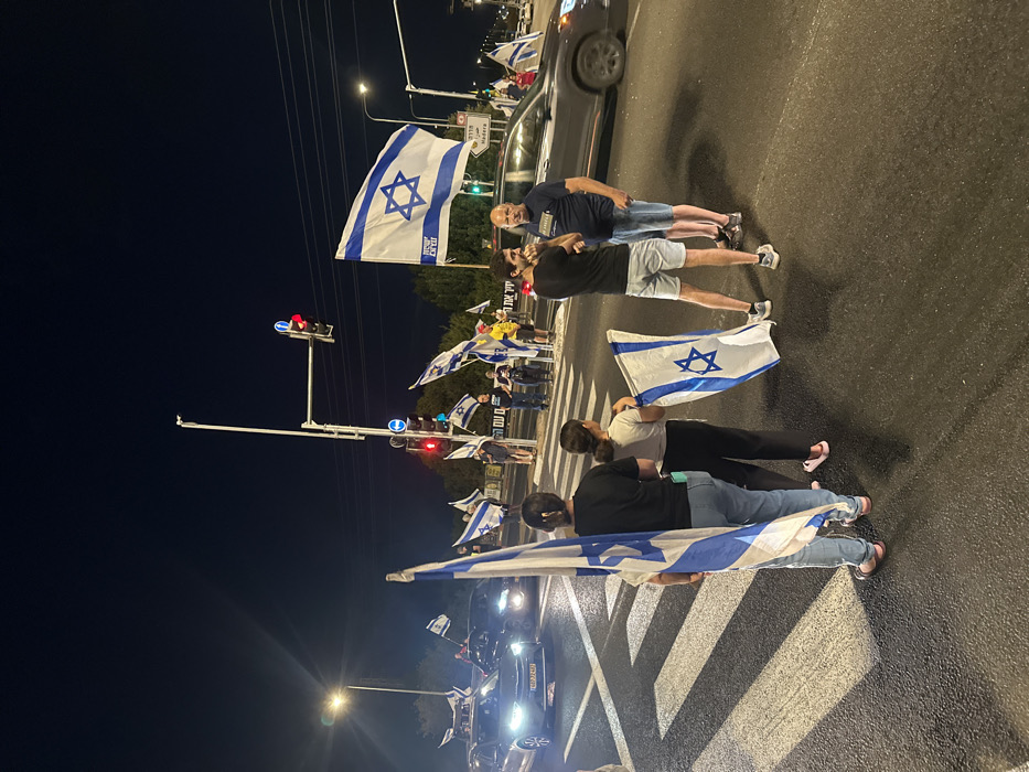 The image displays a nighttime scene at a street intersection in Mishmar Hasharon, Israel. Approximately 10-15 individuals are gathered on and adjacent to a pedestrian crossing and roadway. The sky is dark, illuminated by streetlights, vehicle headlights, and traffic lights, one of which displays a red signal.
In the foreground, a woman with dark hair, wearing a black top and light blue jeans, stands with her back to the camera, holding a large Israeli flag on a pole. Next to her, a younger individual, also facing away, holds a smaller Israeli flag. To their right, a man with a beard, wearing a black t-shirt and light shorts, holds a small Israeli flag. Adjacent to him, a bald man in a dark blue t-shirt and shorts stands, observing the scene.
Multiple other individuals are visible in the mid-ground and background, standing on the sidewalk or near the road edge, predominantly holding Israeli flags of varying sizes. One prominent Israeli flag in the mid-ground, attached to a pole, bears the Hebrew text "עם ישראל חי" (Am Yisrael Chai) below the Star of David. A street sign in the background indicates "Hadera" (חדרה).
At least three vehicles are present on the road: two in the left background with illuminated headlights (one displaying the license plate "848:32:46"), and a gray SUV-like vehicle in the right background, also with illuminated headlights. The road surface is asphalt, featuring white pedestrian crossing stripes. The collective display of Israeli flags suggests a public assembly or demonstration.