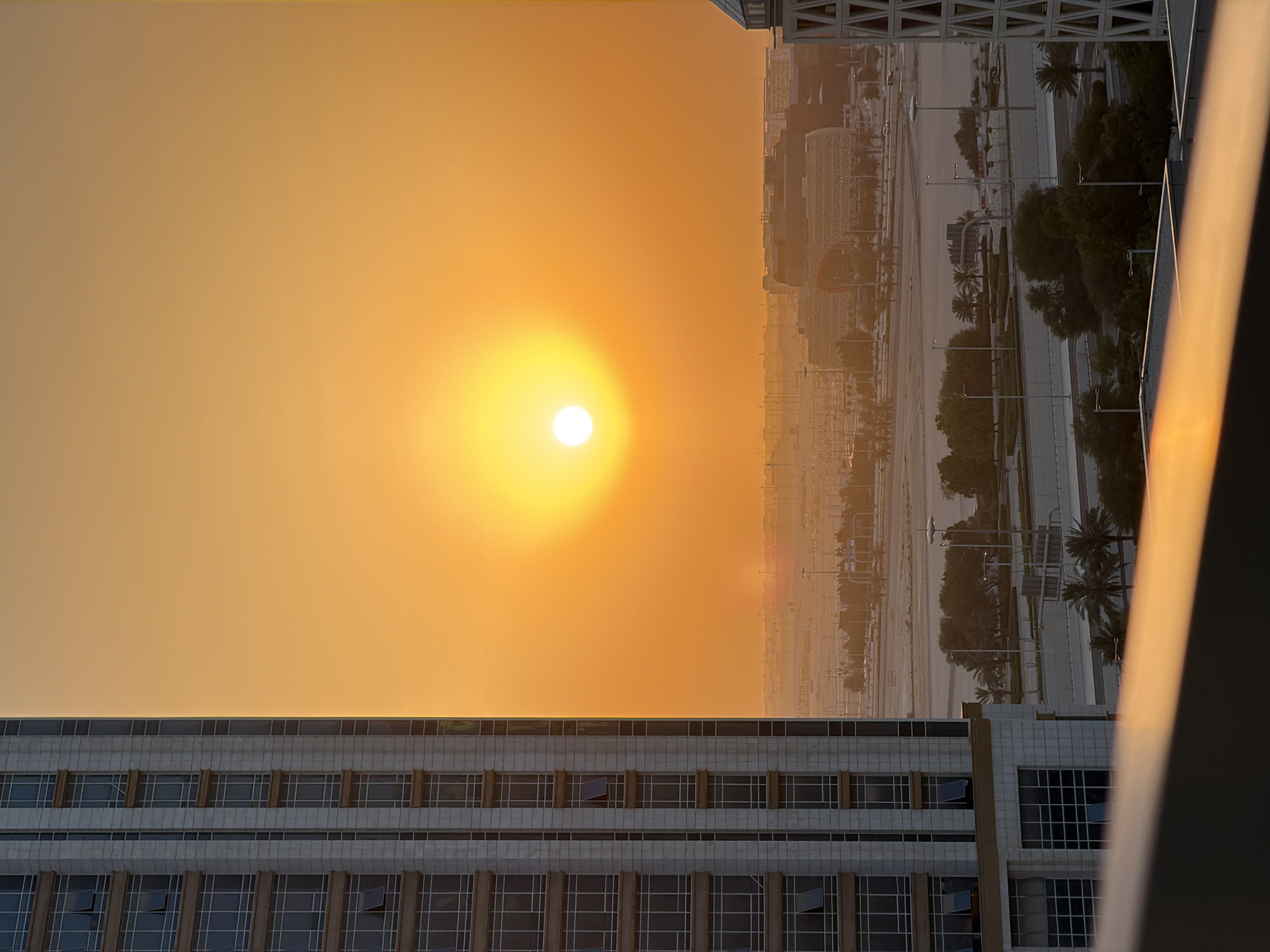 The image depicts a city skyline at sunset, viewed from a high vantage point, possibly a building. The sun, a bright orb, dominates the left side of the frame, casting a warm, orange glow across the sky. The setting sun suggests it is either sunrise or sunset, with the latter being more likely given the term "skyline."
In the foreground, the upper section of a modern building with a facade of windows is visible. Further into the scene, other buildings of varying architectural styles can be seen, creating a dense urban landscape. Trees, including palm trees, dot the landscape, suggesting a warm climate. Roads and pathways are also visible, indicating an active city. The overall impression is one of a serene and beautiful urban environment transitioning into evening. The light quality creates a peaceful and somewhat nostalgic atmosphere. No people are visible in the image, and there is no discernible text.