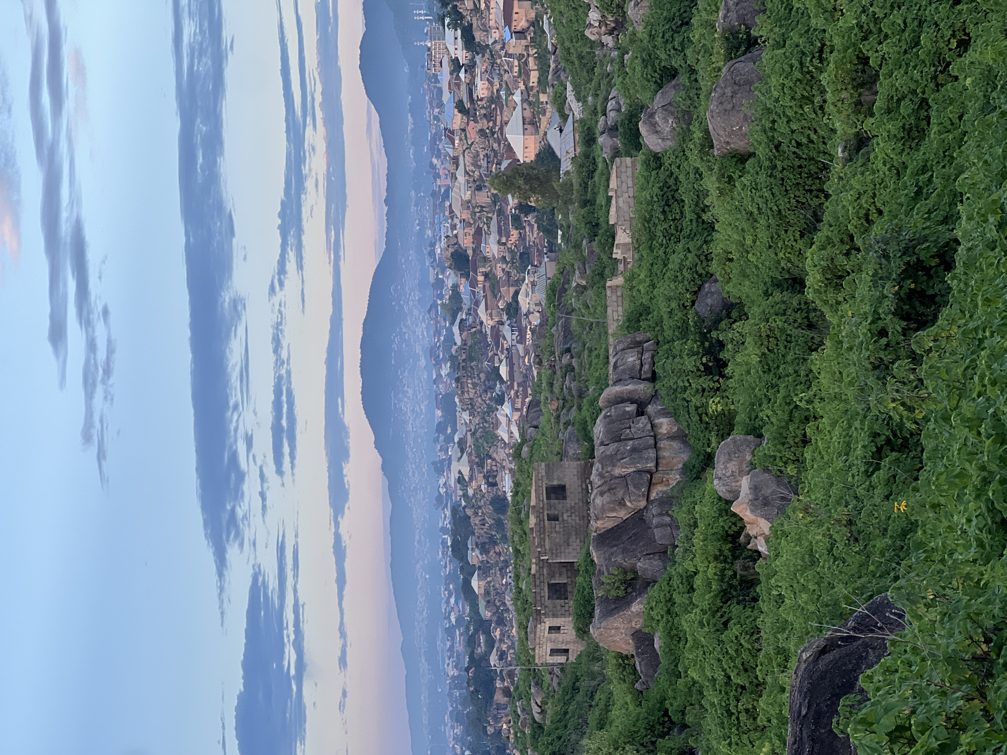 An overhead, medium shot captures the sprawling landscape of Jos, Nigeria, at dawn. A soft, rosy hue from the rising sun paints the sky, which is adorned with wispy, pale blue clouds. In the foreground, a dense canopy of vibrant green foliage and large, weathered grey rocks blankets the terrain. Among them, a partially constructed building made of grey concrete blocks stands as a stark, unfinished structure.
Beyond the immediate rocky and vegetated foreground, the city of Jos unfolds. A dense collection of buildings, characterized by their varied architectural styles and colorful rooftops – predominantly terracotta, beige, and white – sprawls across the landscape. These structures are nestled amongst the natural rock formations that are characteristic of Jos. In the distance, a range of hazy, blue-tinged mountains forms a dramatic backdrop, their peaks softened by the morning mist and the atmospheric haze. The overall scene depicts a harmonious, yet rugged, coexistence between the urban environment and the striking natural topography of Jos.