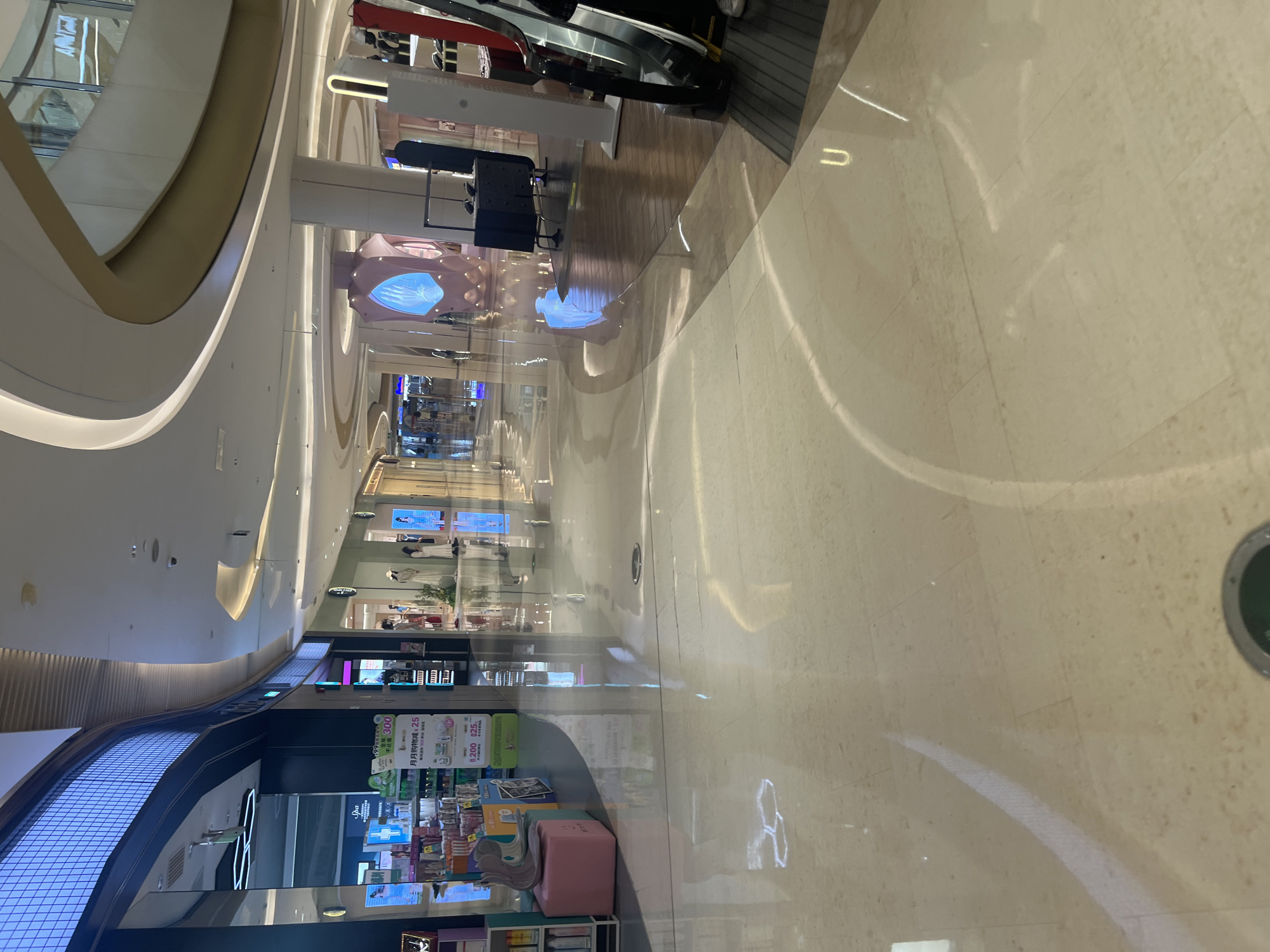 The image shows the interior of a modern shopping mall in Yuzhong District, China. The mall features polished marble floors that reflect the overhead lighting and architectural elements. In the background, there are various shopfronts, escalators, and display areas. One shop on the left displays beauty products with prominent signage. Further back, mannequins are dressed in formal wear in another store. The overall atmosphere is bright and spacious, with curved lines and soft lighting integrated into the ceiling and walls. There are a few people visible in the distance, appearing to be shoppers. The environment suggests a high-end retail setting.