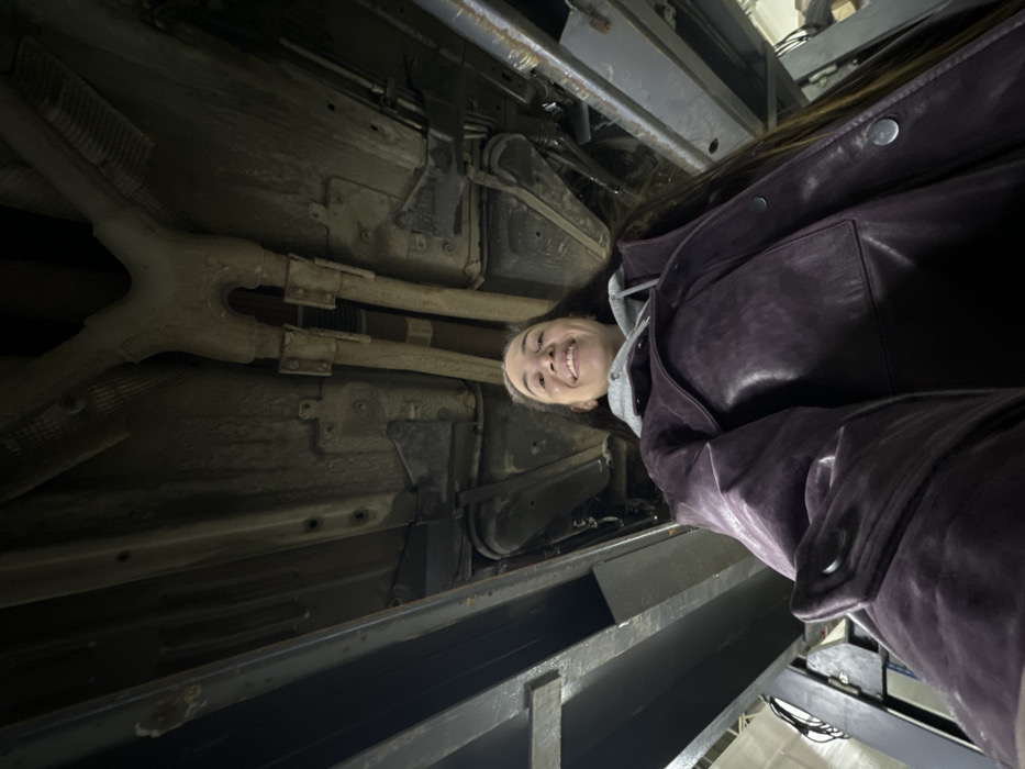 The image depicts a young adult female positioned directly beneath the underside of a raised vehicle within what appears to be an automotive workshop in the city of Zhabkino, Russia. The female, with long dark hair, is smiling broadly and looking upwards towards the camera, suggesting a low-angle selfie. She is wearing a grey hooded sweatshirt under a dark purple, possibly leather, collared jacket with snap buttons. Above her, the vehicle's underside is visible, showcasing elements such as the exhaust system with a Y-pipe and heat-shielded sections, chassis rails, and other components, all appearing dusty and used. On both the left and right sides of the frame, robust metal support structures of what is identified as an automotive lift are prominently displayed, holding the vehicle in an elevated position. No other individuals are visible.