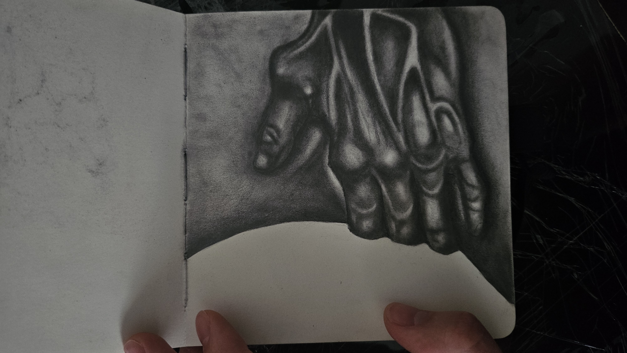 A charcoal drawing on paper depicts a close-up of a hand in motion, rendered with strong chiaroscuro to emphasize its form and texture. The hand is foreshortened and positioned in the lower right quadrant of the image, with fingers curled and appearing to grasp something unseen. The shading creates a dramatic contrast between light and shadow, highlighting the musculature and bone structure of the hand. The background is subtly textured, suggesting a neutral environment that doesn't detract from the subject. To the left of the hand, a curved, lighter area suggests the edge of another object or surface, possibly part of the same composition or an adjacent page in a sketchbook. The drawing is bound by stitching along the left side, indicating it is part of an open sketchbook. The presence of fingers holding the page suggests the drawing is being viewed or presented. The overall impression is one of artistic study and skillful rendering of human anatomy.