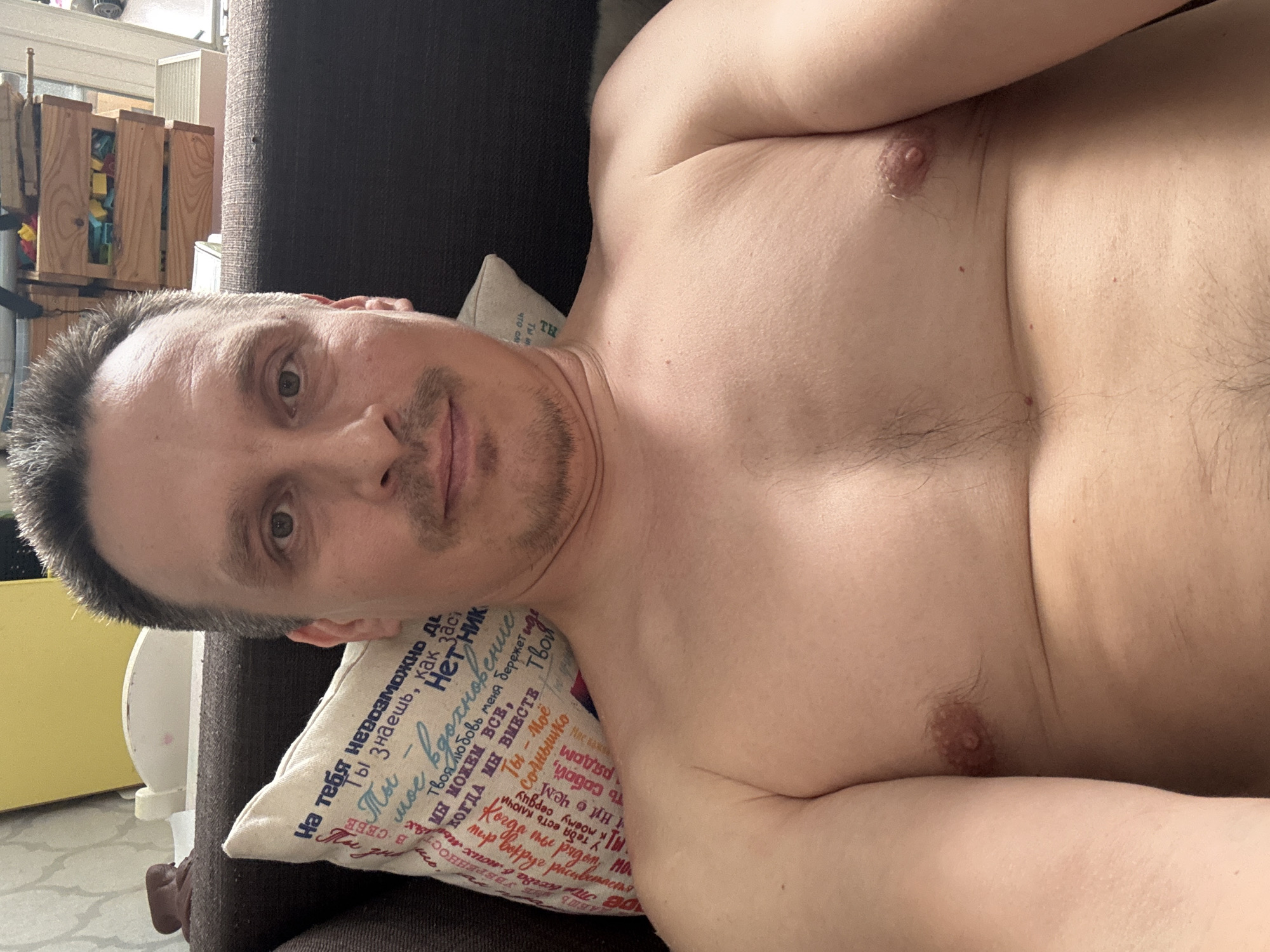 A middle-aged Caucasian man with short graying hair and a mustache is reclining on a dark-colored couch. He is shirtless, with visible chest hair. He is resting his head on a pillow with text in Russian on it. In the background, there's a wooden shelving unit and what appears to be a yellow-painted object. The city of Izhevsk, Russia, is not discernible in the image.