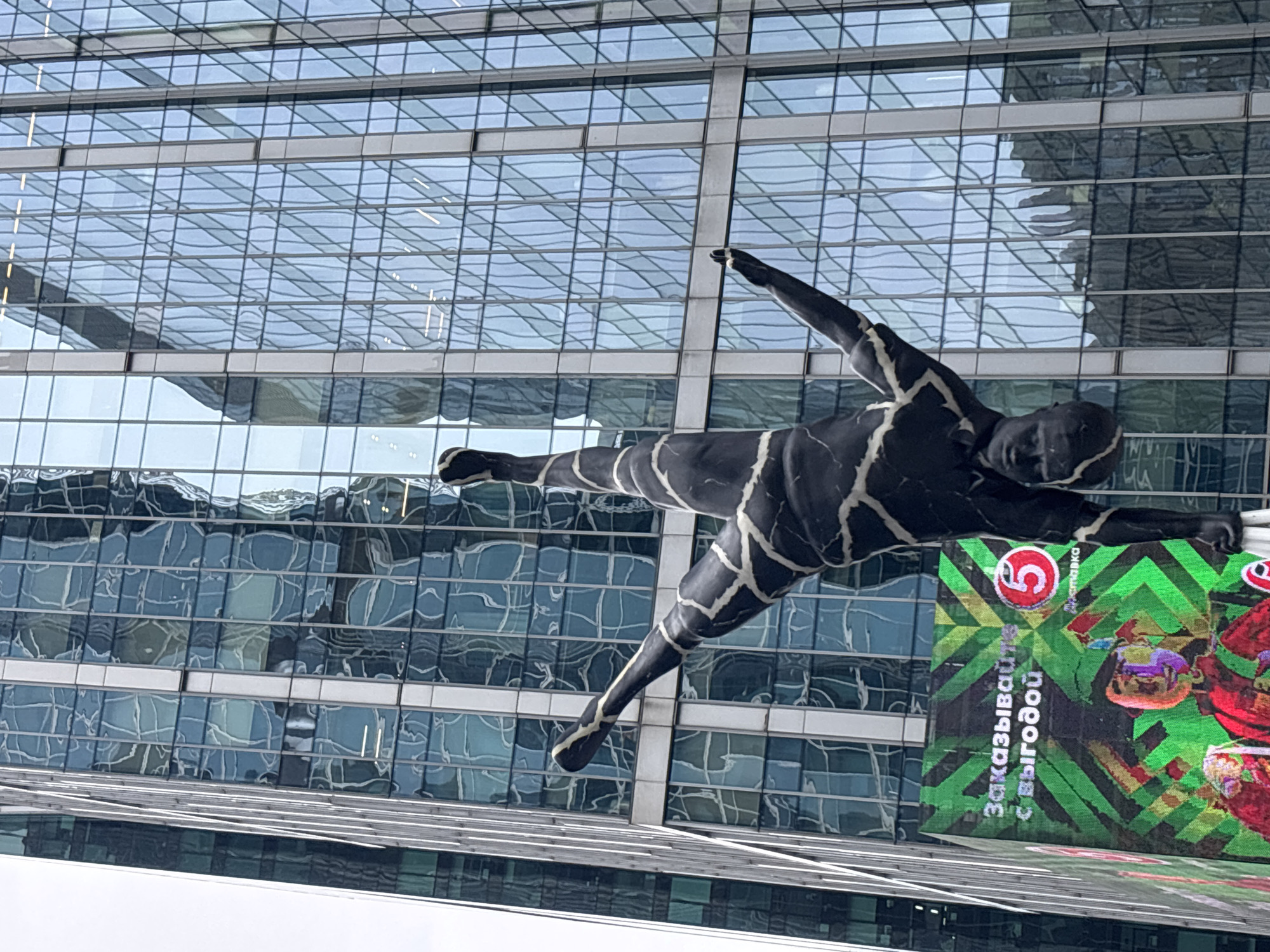 The image depicts a sculpture of a gymnast in front of a modern glass building in Moscow, Russia. The statue is dark gray with white markings resembling cracks. The gymnast is posed mid-air, with one arm extended forward and the other back. A large sign, with the logo of a Russian grocery chain, "Pyaterochka" (a "5" symbol), is next to the statue. The background building has a grid pattern of reflective glass windows. The sky is overcast.