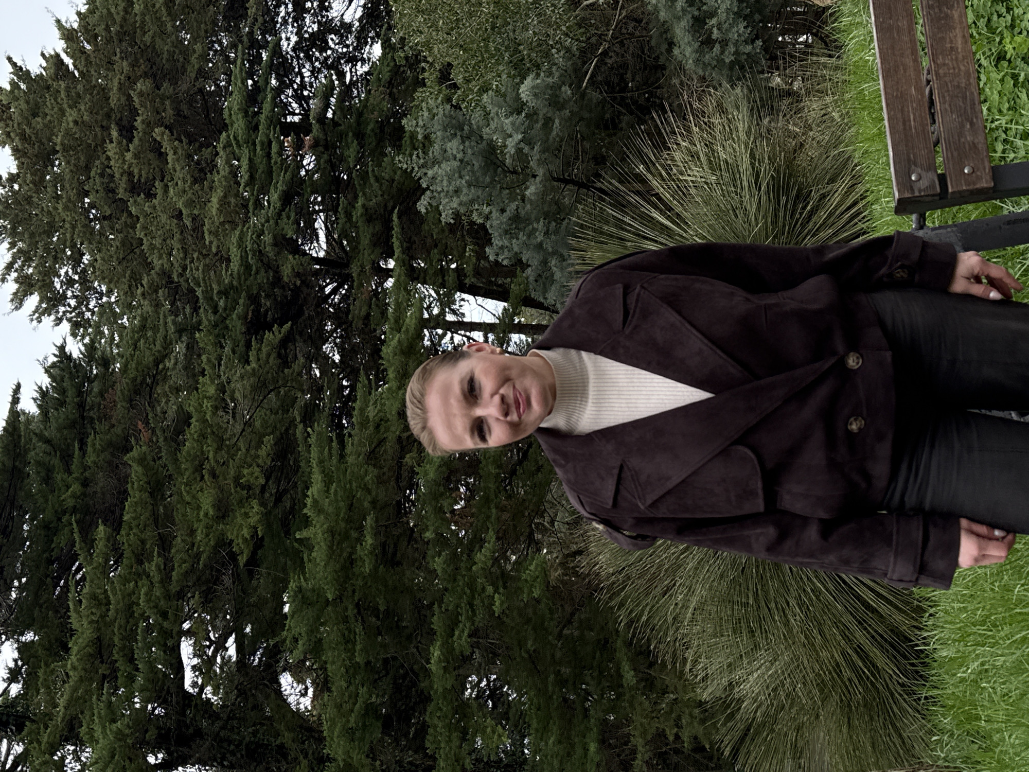 The image displays a female subject standing outdoors in a park or garden environment within the city of Sochi, Russia. The individual, positioned centrally, faces the camera directly with a subtle smile. She has light-colored hair pulled back. She is dressed in a cream-colored turtleneck sweater layered under a dark brown, textured jacket featuring wide lapels, two visible buttons on the front closure, and strap details on the cuffs. She wears dark-colored trousers. The background consists predominantly of dense, dark green coniferous trees, characteristic of Sochi's subtropical vegetation. To the subject's left, a large shrub with spiky, linear green foliage is visible. To her right, a tree with lighter, silvery-green leaves can be seen. The ground is covered with green grass, and a portion of a wooden park bench with dark metal supports is present in the lower right foreground. The scene is illuminated by diffuse, natural light, suggesting an overcast sky.