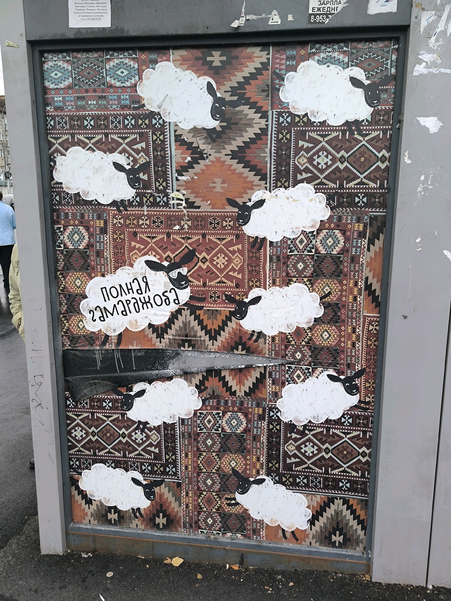 The image depicts a close-up, eye-level view of a portion of a weathered, outdoor advertisement panel, likely a bus stop shelter, situated in Novosibirsk, Russia. The panel features a prominent, colorful, patterned textile as its primary background, resembling a traditional rug with geometric designs in shades of red, brown, blue, and orange. Affixed to this backdrop are several cartoonish depictions of white sheep with black heads and ears, some appearing to be jumping or running across the pattern.
One sheep, centrally located and slightly below the middle of the panel, is enclosed within a cloud-shaped sticker that has text written in Russian. The text, rendered in a playful, hand-drawn font, appears to read "Полная замарагожа" which translates to "complete mess" or "utter chaos." This sticker also includes a caricature of a sheep's head wearing sunglasses.
Other sheep are scattered across the panel, positioned in various locations and orientations, contributing to a whimsical and somewhat disheveled aesthetic. The advertisement panel itself shows signs of age and wear, with some peeling edges and tape visible. A section near the bottom of the panel appears to be damaged or covered with a dark, reflective material.
The environment suggests an urban setting, with the panel mounted on a gray, metallic frame. The ground in front of the panel is visible and consists of asphalt and loose debris. To the left of the panel, a sliver of a person wearing a light blue shirt can be seen, along with a glimpse of a yellow jacket. The overall impression is that of public art or a humorous sticker campaign applied to an advertising space in a city.