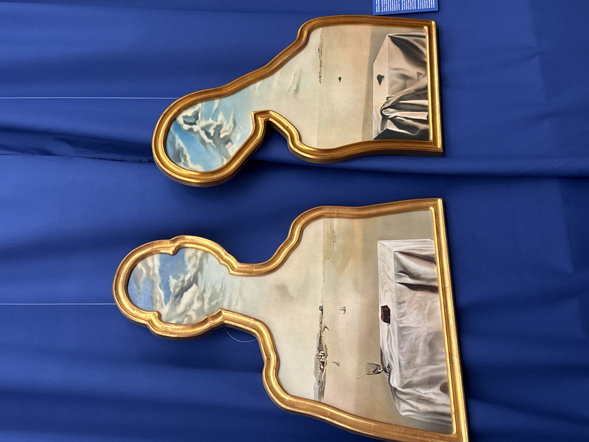The image displays two silhouette-shaped paintings, framed in gold, hanging against a vibrant blue curtain. The paintings, which appear to be in the surrealist style of Salvador Dalí, depict dreamlike landscapes within the contours of human forms.
The left painting showcases a coastal town or city in the distance, with a table covered by a white cloth in the foreground. On the table, a glass with a straw and a small rectangular object, possibly a piece of chocolate, are visible. The upper portion of this silhouette is filled with a sky filled with dramatic clouds.
The right painting, positioned slightly lower and tilted, features a similar surreal landscape. A table draped with a wrinkled white cloth is present, this time with a cluster of dark objects, perhaps grapes, on it. A solitary dark shape floats in the distant landscape. The upper part of this silhouette also contains a sky with clouds.
The blue curtain behind the paintings suggests an indoor exhibition space. A small piece of paper with text is visible on the right side of the image, attached to the curtain.
Based on external information, these paintings are part of an exhibition of Salvador Dalí's works at the Daria Cultural Center (Daria Art Gallery) in Pernik, Bulgaria. The exhibition, titled "The Persistence of Memory," features holographic reproductions and original graphic works by the artist.
