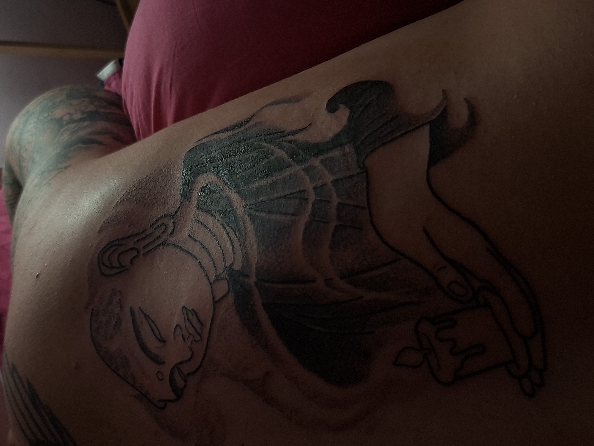 A tattoo of a woman's face holding a candle is on the back. There is another tattoo on the shoulder. A pink pillow is in the upper background. The location is not Pleven, Bulgaria.