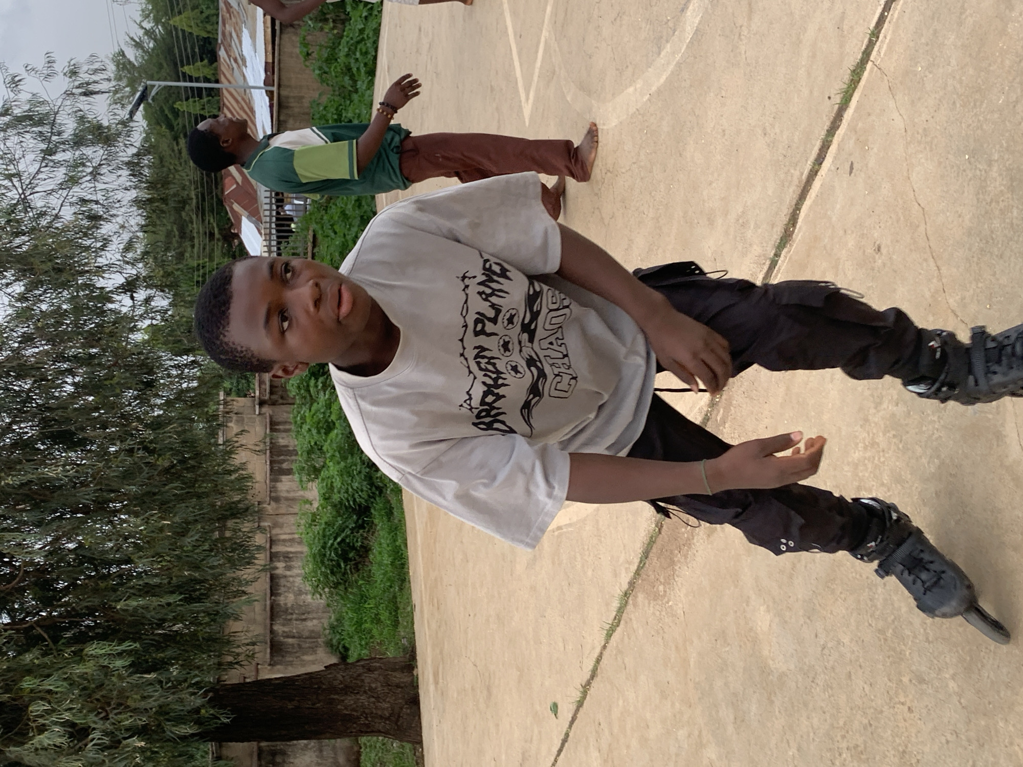 The image shows a young Black man wearing a grey t-shirt with a graphic design, black cargo pants, and black inline skates. He stands on a concrete surface with white lines that suggest a sports court, possibly a basketball or volleyball court. He faces the camera with a neutral expression. Another person stands barefoot behind him, partially obscured. The background features lush greenery, a tall tree, and a fence. In the distance are buildings with corrugated metal roofs, typical of urban areas in Jos, Nigeria. The setting appears to be outdoors during daylight hours.