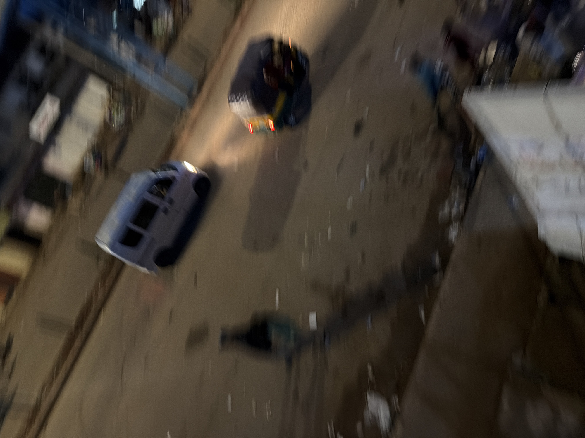 The media file captures a night scene in Bukuru, Nigeria, with a notable blur indicating movement, likely from a moving vehicle or camera shake. Two vehicles are visible on a road. The first, a light-colored van, is positioned in the foreground, its headlights illuminated. Behind it, a darker vehicle, possibly a car or bus, is also visible, with its taillights glowing red. The surrounding environment appears to be an urban or semi-urban area, with some structures and lighting visible in the background, suggesting a commercial or residential district. The ground surface is paved, and there are scattered debris, hinting at possibly untidy conditions or recent activity. The time of day is clearly night, indicated by the darkness and the artificial lights. No specific weather conditions are discernible due to the blur and lighting. The overall impression is a snapshot of urban life at night, with vehicles in motion. There is no visible text in the media file.